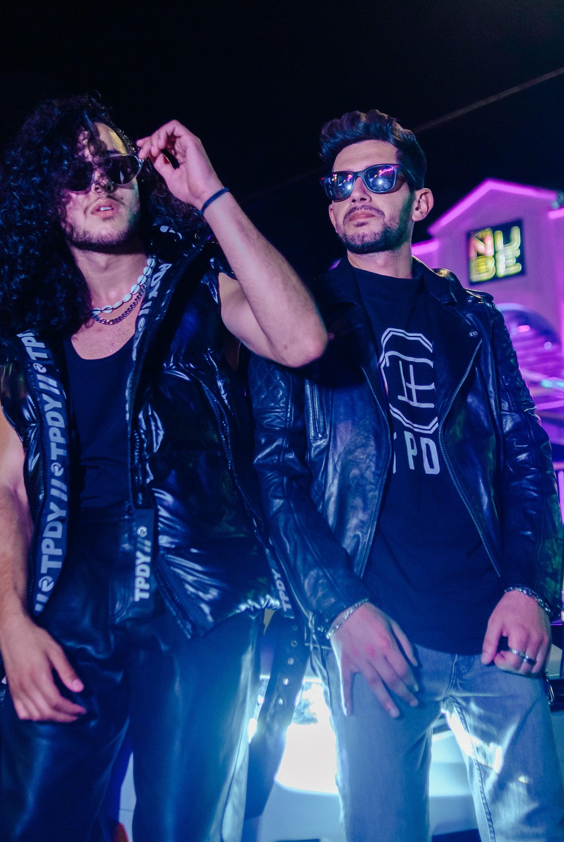 Zwei Männer mit modischer Lederjacke und Weste vor einem Cabrio. Im Hintergrund eine Diskothek bei Nacht