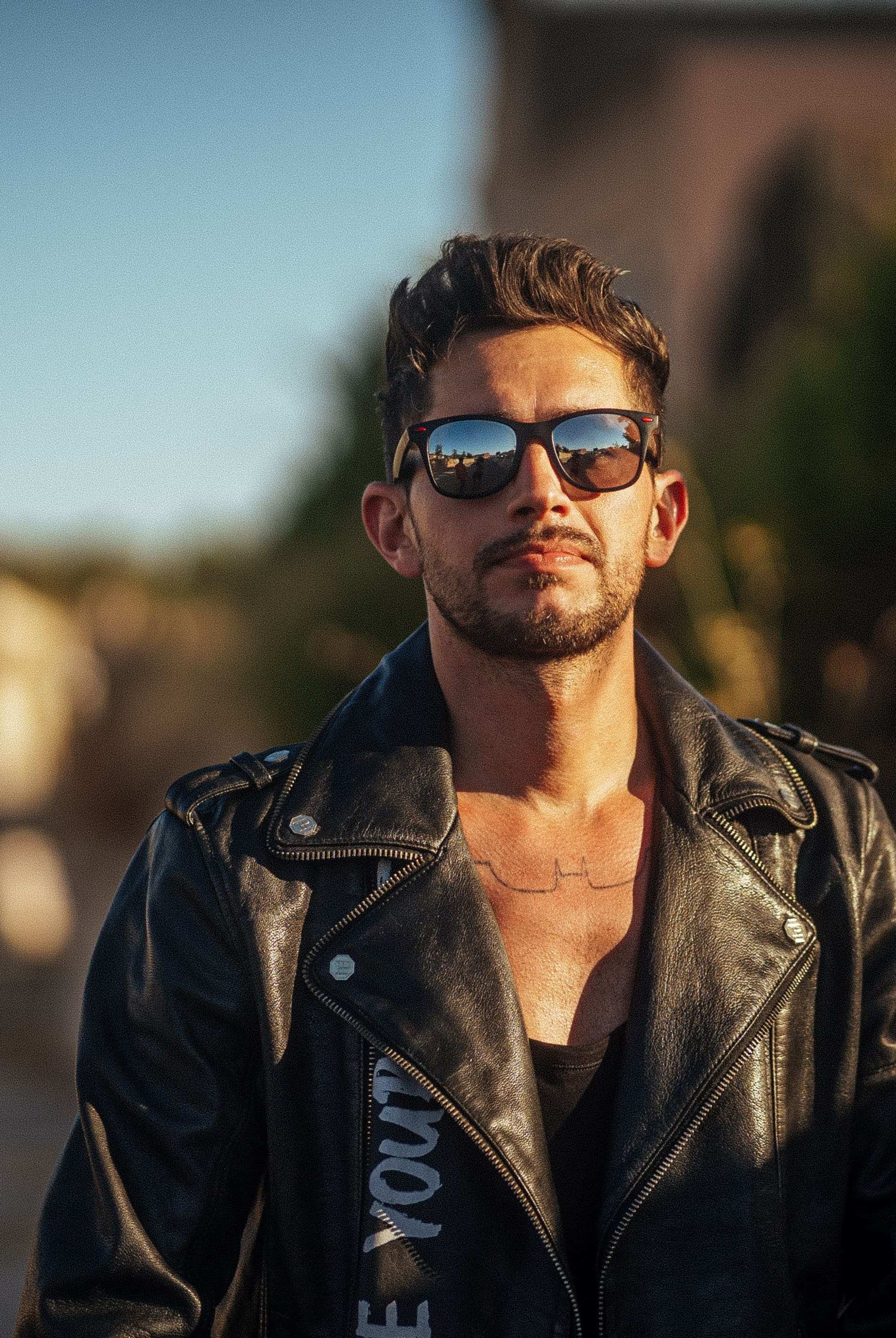 Ein Mann in schwarzer Lederjacke und Sonnenbrille steht im Freien und blickt in die Kamera. Der Hintergrund wirkt sonnig und unscharf, man sieht Grünflächen und Gebäude.