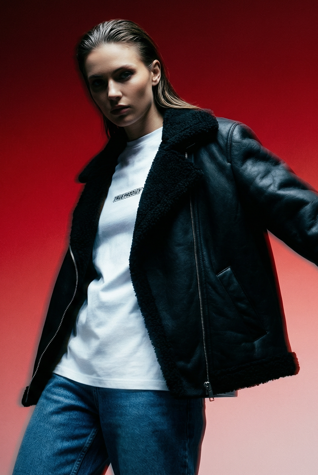 Eine Frau mit blonden Haaren, einer schwarzen Lederjacke, einem schwarzen Outfit und schwarzen Stiefeln sitzt auf dem Boden vor einer weißen Backsteinwand und berührt mit einer Hand ihren Kopf. Sonnenlicht wirft Schatten an die Wand.