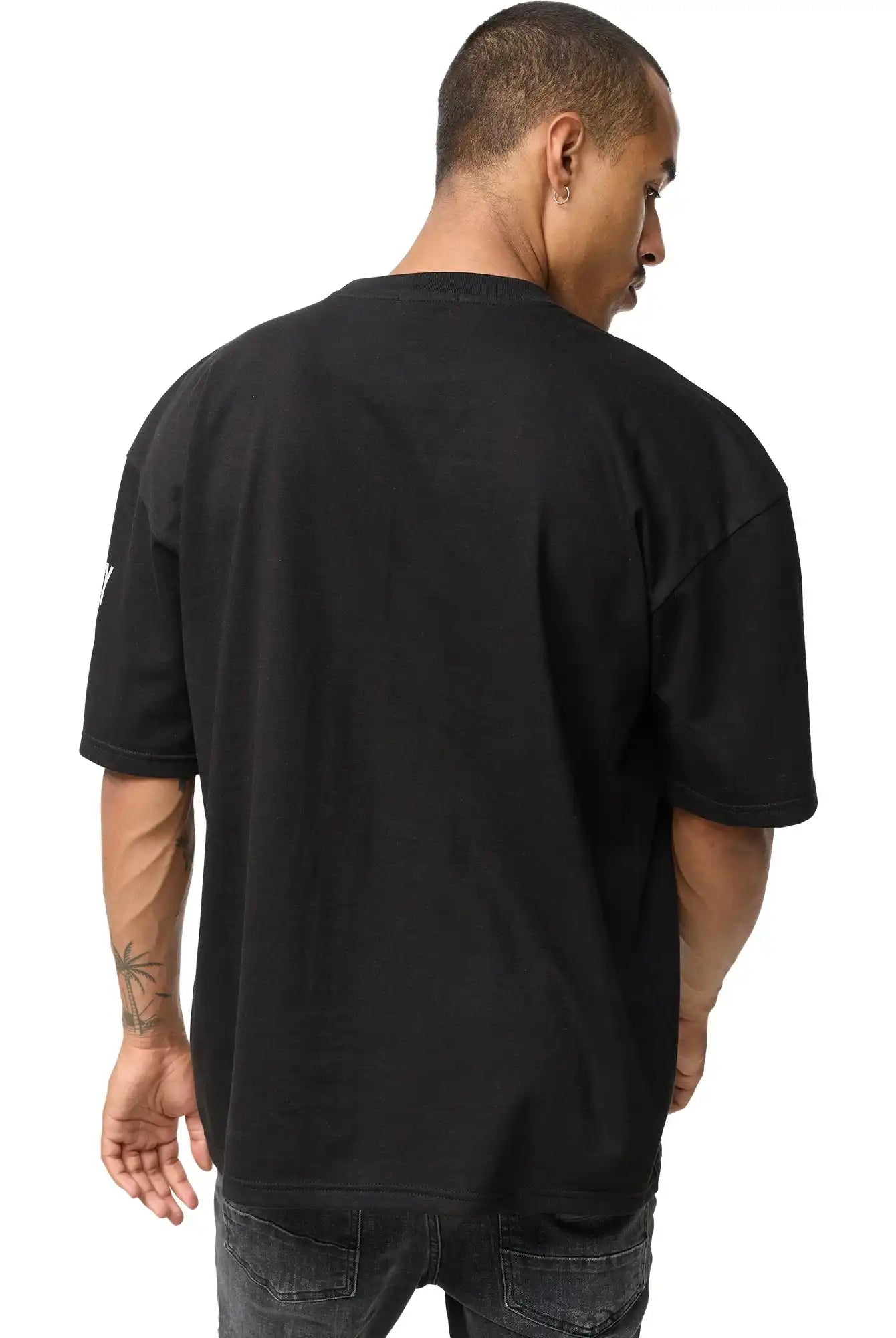 Eine Person mit kurzen Haaren und einer Tätowierung auf dem linken Unterarm ist von hinten zu sehen. Sie trägt das Adrian Oversized T-Shirt mit Druck von trueprodigy und dunkle Jeans und steht vor einem schlichten weißen Hintergrund.