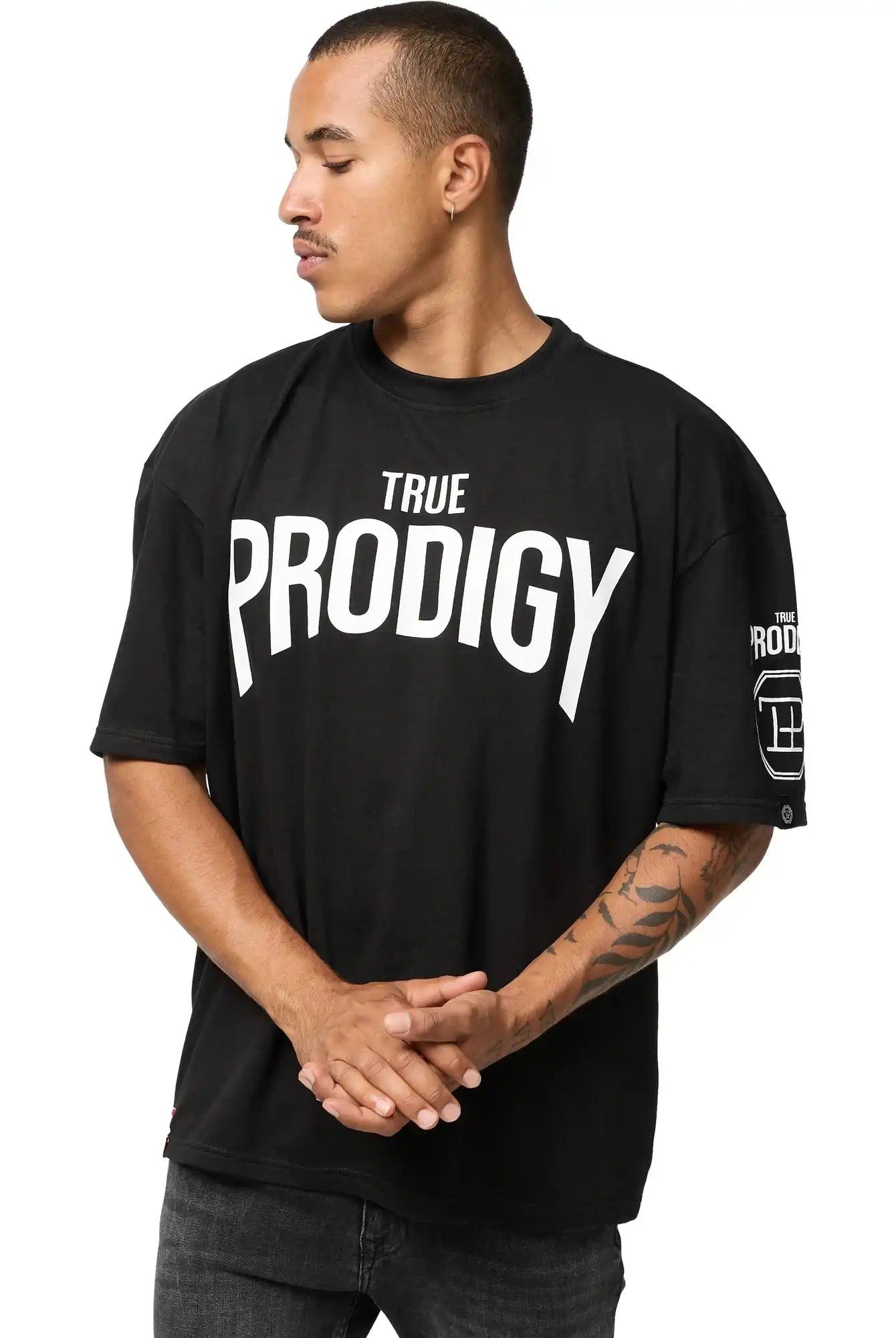 Auf dem Adrian Oversized T-Shirt mit Druck von trueprodigy in schwarzem Heavy Jersey mit fettem weißen "TRUE PRODIGY"-Schriftzug steht eine Person seitlich mit gefalteten Händen und zeigt ein Tattoo auf dem linken Unterarm.
