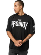 Auf dem Adrian Oversized T-Shirt mit Druck von trueprodigy in schwarzem Heavy Jersey mit fettem weißen "TRUE PRODIGY"-Schriftzug steht eine Person seitlich mit gefalteten Händen und zeigt ein Tattoo auf dem linken Unterarm.