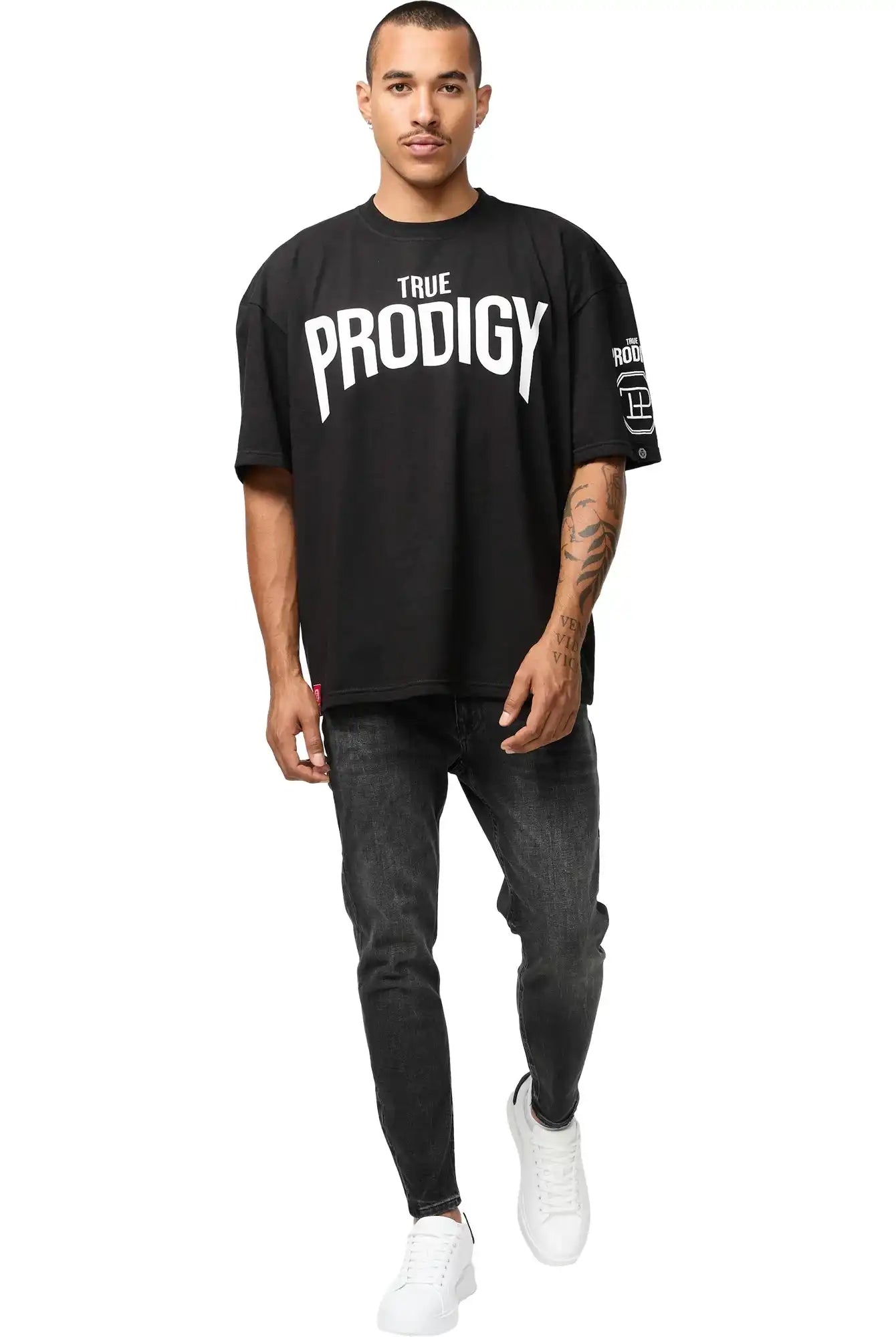 Ein Mann mit kurzen Haaren und Schnurrbart trägt das Adrian Oversized T-Shirt mit Druck von trueprodigy in schwarz, gepaart mit schwarzen Skinny Jeans und weißen Turnschuhen, und geht auf die Kamera vor einem weißen Hintergrund zu.