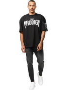 Ein Mann mit kurzen Haaren und Schnurrbart trägt das Adrian Oversized T-Shirt mit Druck von trueprodigy in schwarz, gepaart mit schwarzen Skinny Jeans und weißen Turnschuhen, und geht auf die Kamera vor einem weißen Hintergrund zu.