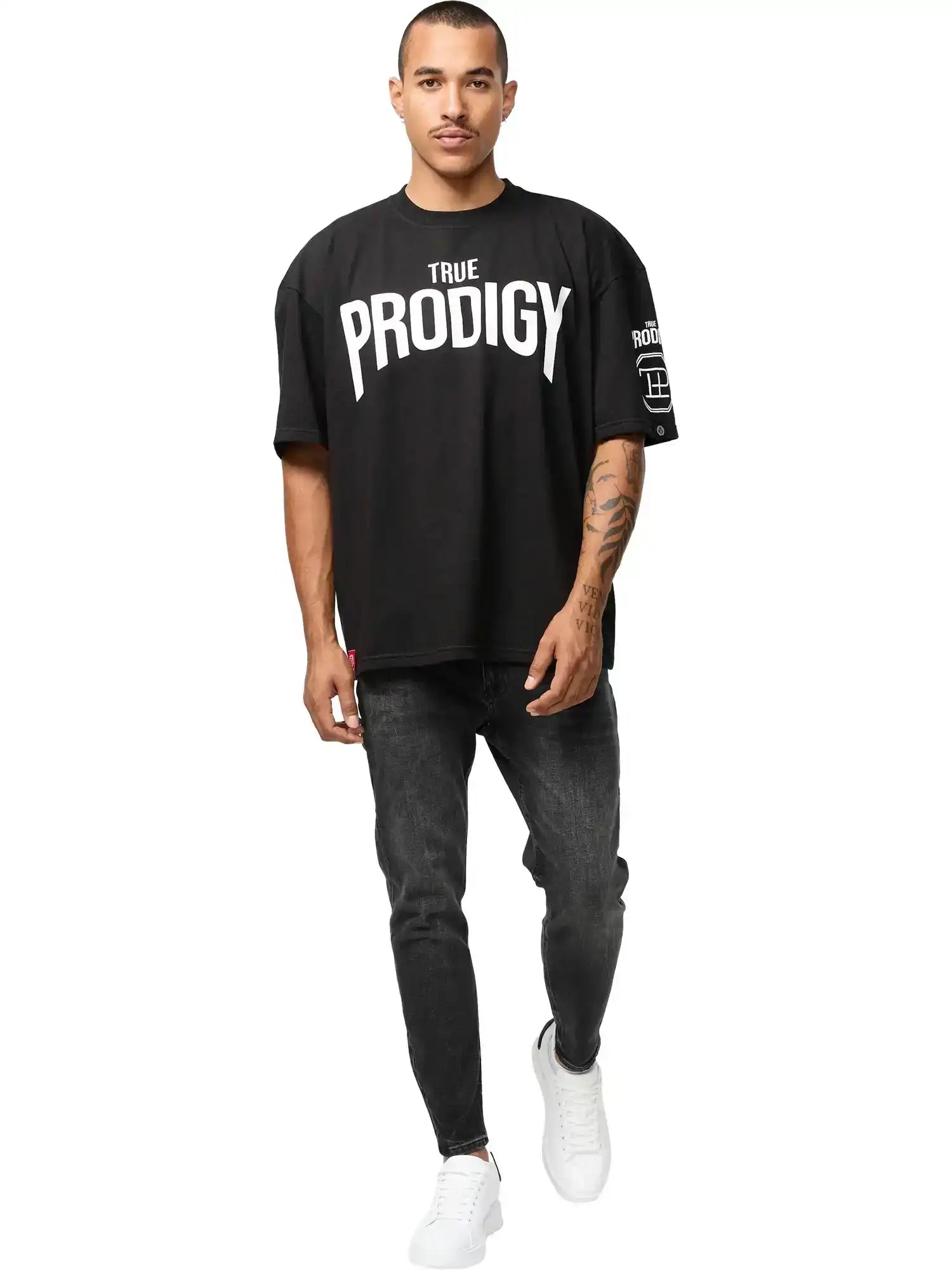 Ein Mann mit kurzen Haaren und Schnurrbart trägt das Adrian Oversized T-Shirt mit Druck von trueprodigy in schwarz, gepaart mit schwarzen Skinny Jeans und weißen Turnschuhen, und geht auf die Kamera vor einem weißen Hintergrund zu.