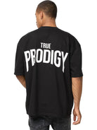 Eine Person blickt von der Kamera weg und trägt das trueprodigy Jona Oversized T-Shirt mit Druck in schwarz mit dem Aufdruck TRUE PRODIGY in fetten weißen Buchstaben auf dem Rücken, gepaart mit einer dunklen Jeans.