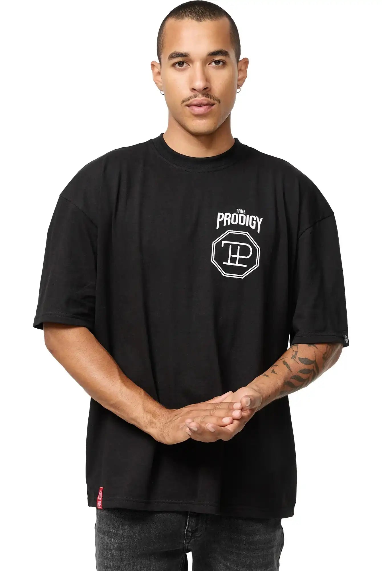 Eine Person mit Kurzhaarschnitt und Schnurrbart steht mit dem Gesicht nach vorne, trägt das Jona Oversized T-Shirt mit Druck von trueprodigy, mit lockerer Passform und weißem "The Prodigy"-Logo auf der linken Brust, die Hände vor sich verschränkt.