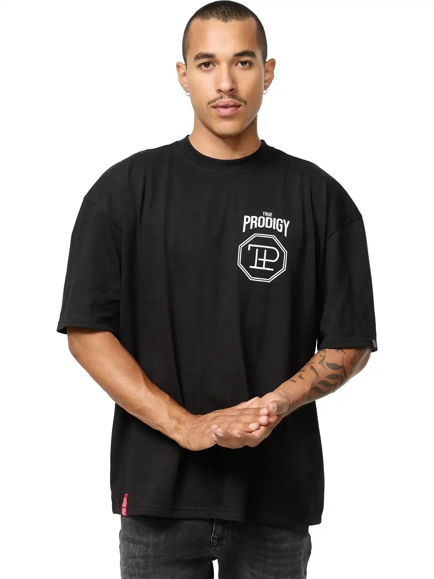 Eine Person mit Kurzhaarschnitt und Schnurrbart steht mit dem Gesicht nach vorne, trägt das Jona Oversized T-Shirt mit Druck von trueprodigy, mit lockerer Passform und weißem "The Prodigy"-Logo auf der linken Brust, die Hände vor sich verschränkt.