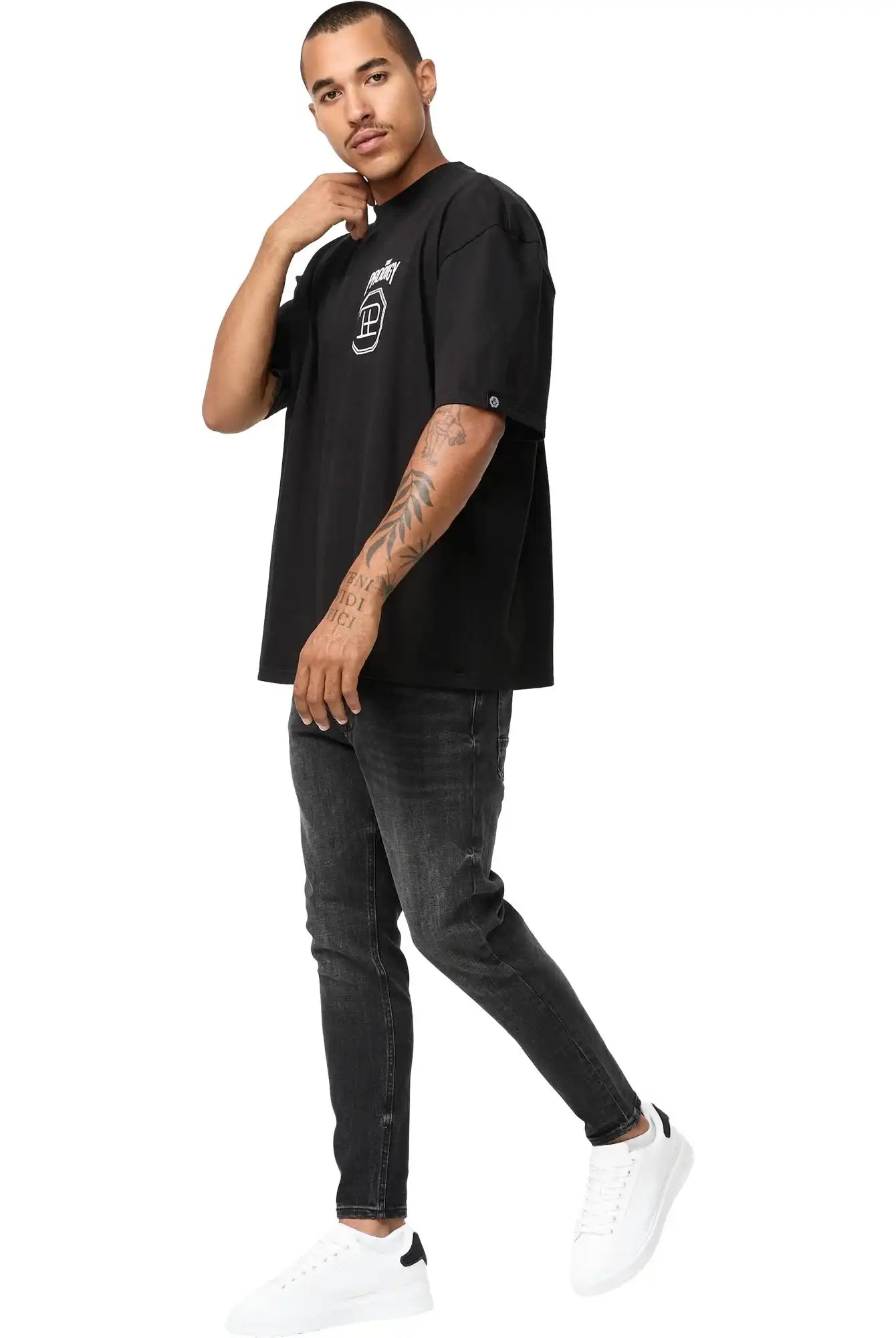 Ein Mann mit Tattoos auf dem linken Arm steht vor einem weißen Hintergrund und trägt das trueprodigy Jona Oversized T-Shirt mit Druck in schwarz, gepaart mit einer schwarzen Jeans und weißen Sneakers. Er blickt leicht nach links.