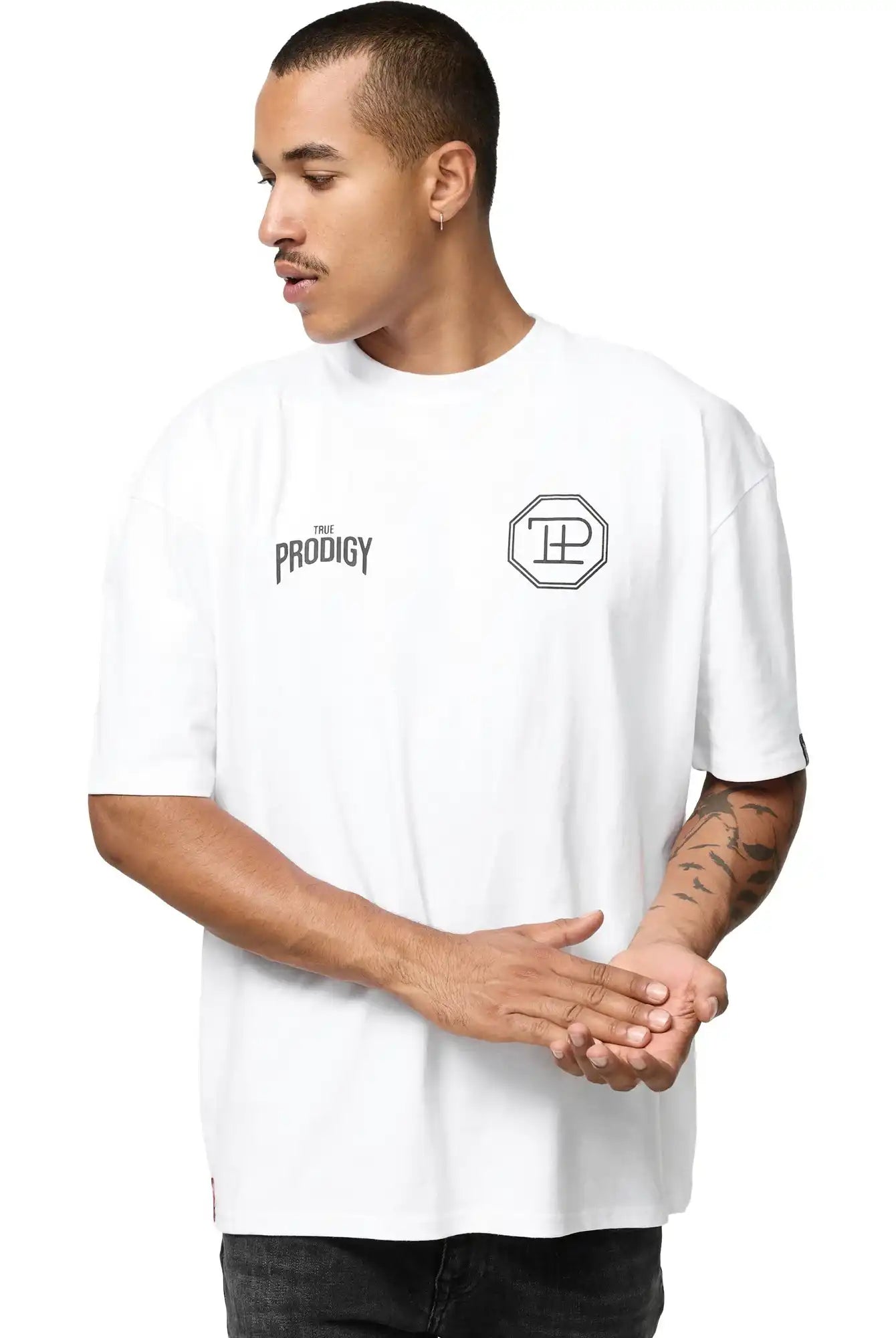 Eine Person trägt das Robin Oversized T-Shirt mit Druck von trueprodigy, ein weißes Unisex-Shirt mit THE PRODIGY-Schriftzug und einem achteckigen Logo auf der Vorderseite, steht mit zusammengelegten Händen und schaut zur Seite.