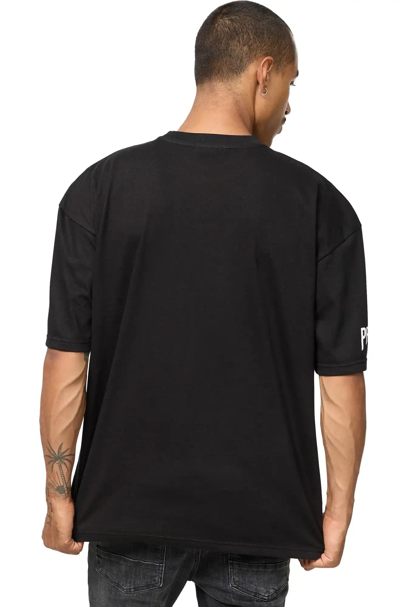 Eine Person mit kurzen Haaren und sichtbaren Tattoos am rechten Unterarm steht mit dem Rücken zur Kamera. Sie trägt das trueprodigy Dilan Oversized T-Shirt mit Druck und verwaschene schwarze Jeans.