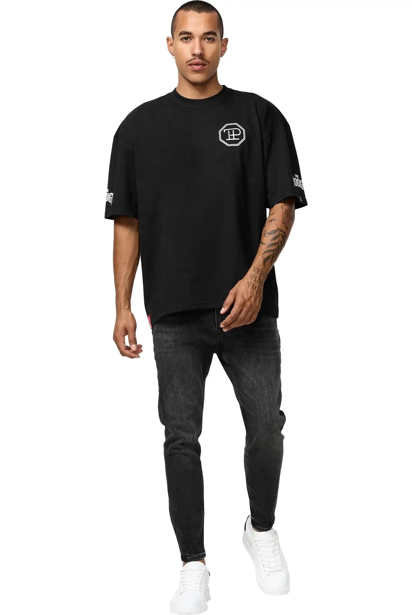 Ein Mann mit kurzen Haaren, gestutztem Schnurrbart und Tattoos am linken Unterarm posiert vor einem weißen Hintergrund und trägt das trueprodigy Dilan Oversized T-Shirt mit Druck, gepaart mit einer schwarzen Jeans und weißen Turnschuhen.
