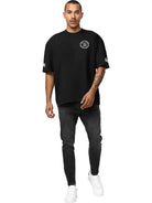 Ein Mann mit kurzen Haaren, gestutztem Schnurrbart und Tattoos am linken Unterarm posiert vor einem weißen Hintergrund und trägt das trueprodigy Dilan Oversized T-Shirt mit Druck, gepaart mit einer schwarzen Jeans und weißen Turnschuhen.