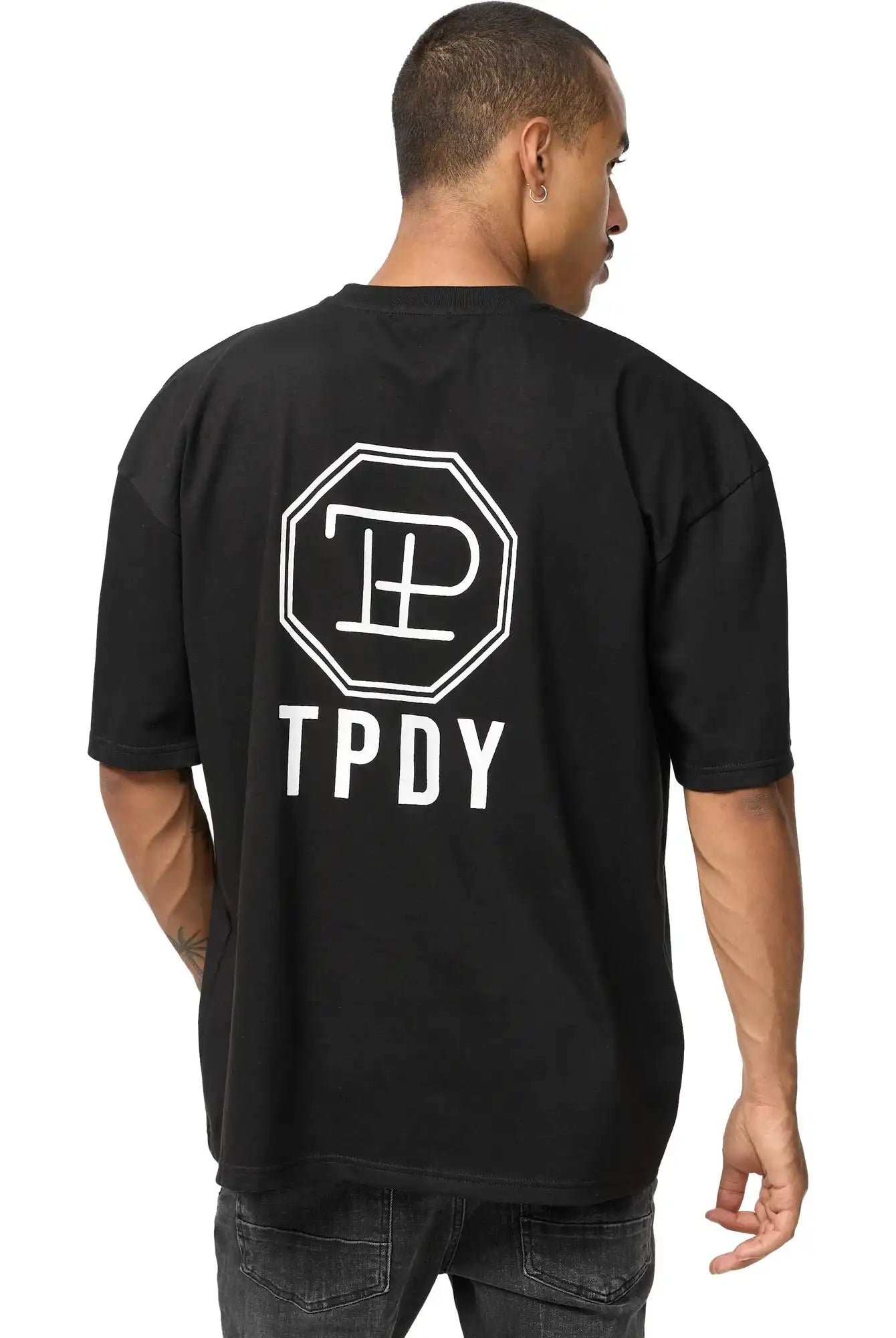 Eine Person blickt von sich weg und trägt das Maxime Oversized T-Shirt mit Druck von trueprodigy - ein schwarzes Unisex-T-Shirt aus schwerem Jersey mit einem weißen, achteckigen Logo und "TPDY" auf dem Rücken - zu einer dunklen Jeans, einen Arm leicht angewinkelt.