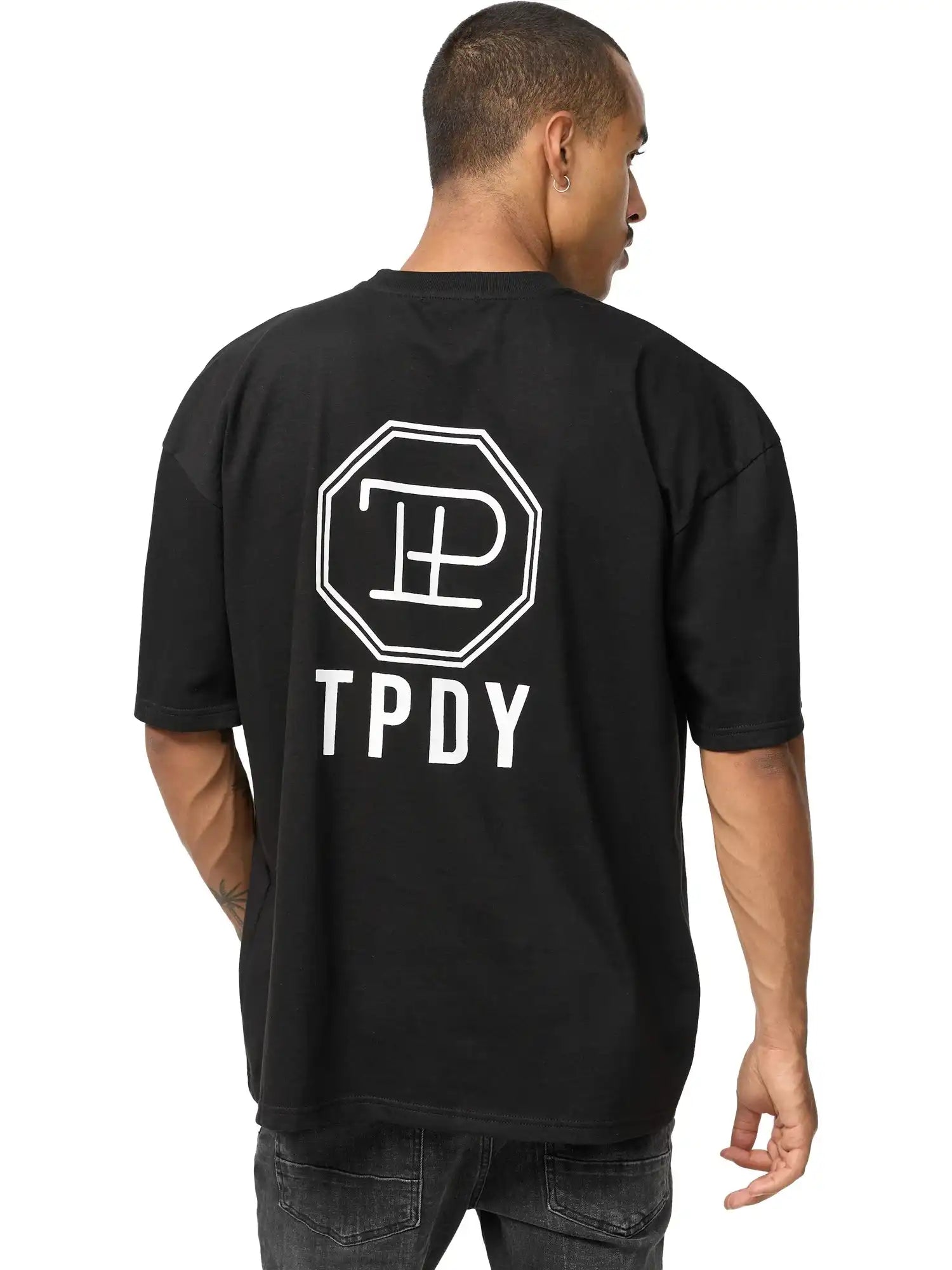 Eine Person blickt von sich weg und trägt das Maxime Oversized T-Shirt mit Druck von trueprodigy - ein schwarzes Unisex-T-Shirt aus schwerem Jersey mit einem weißen, achteckigen Logo und "TPDY" auf dem Rücken - zu einer dunklen Jeans, einen Arm leicht angewinkelt.