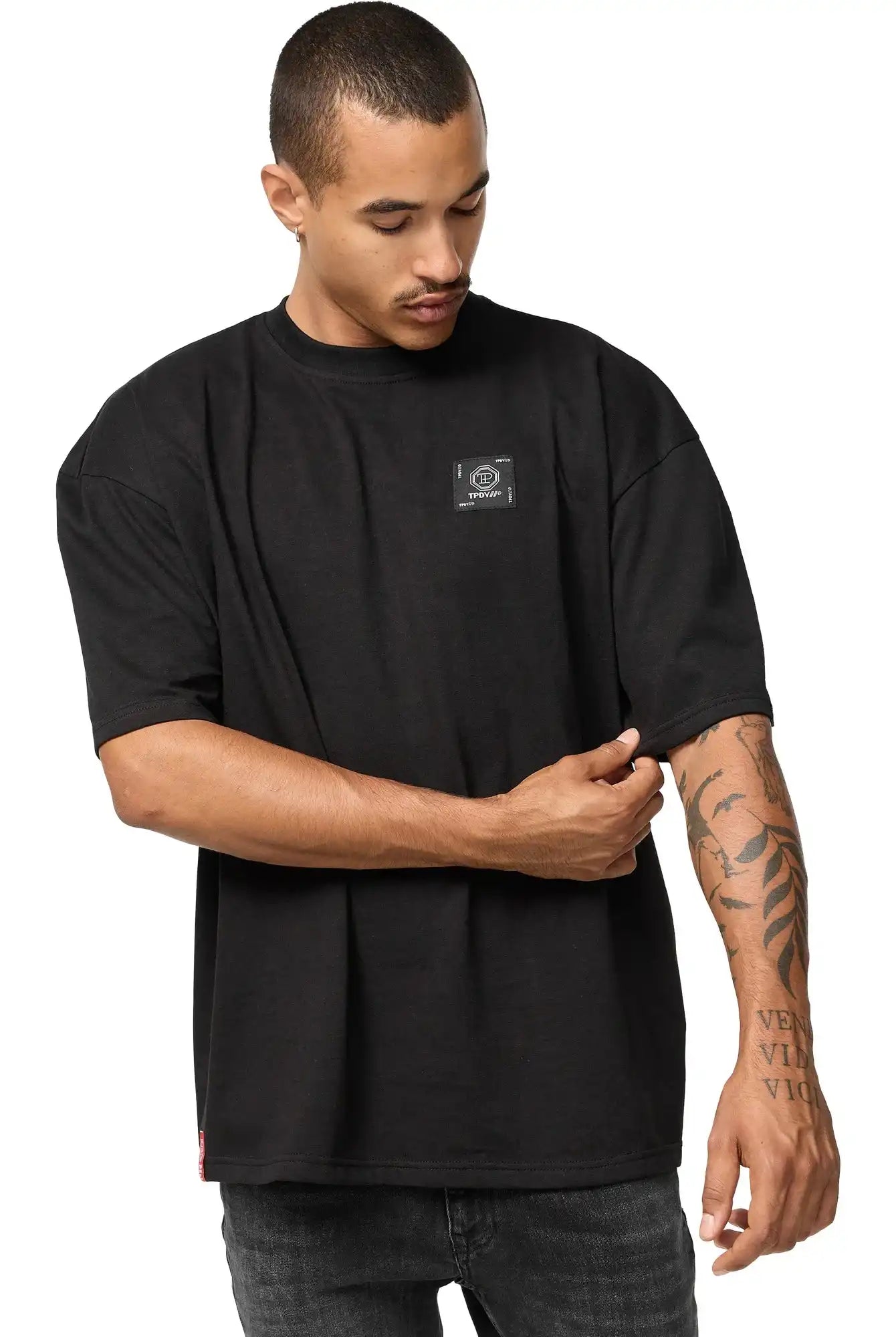 Ein Mann trägt das trueprodigy Maxime Oversized T-Shirt mit Druck in schwarz, mit einem kleinen rechteckigen Logo auf der Brust. Er steht vor einem weißen Hintergrund, rückt seinen Ärmel zurecht und blickt nach unten, wo er Tätowierungen auf seinem Unterarm sieht.