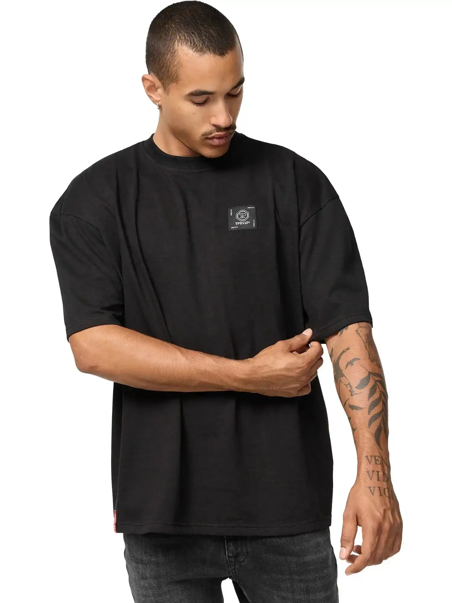 Ein Mann trägt das trueprodigy Maxime Oversized T-Shirt mit Druck in schwarz, mit einem kleinen rechteckigen Logo auf der Brust. Er steht vor einem weißen Hintergrund, rückt seinen Ärmel zurecht und blickt nach unten, wo er Tätowierungen auf seinem Unterarm sieht.