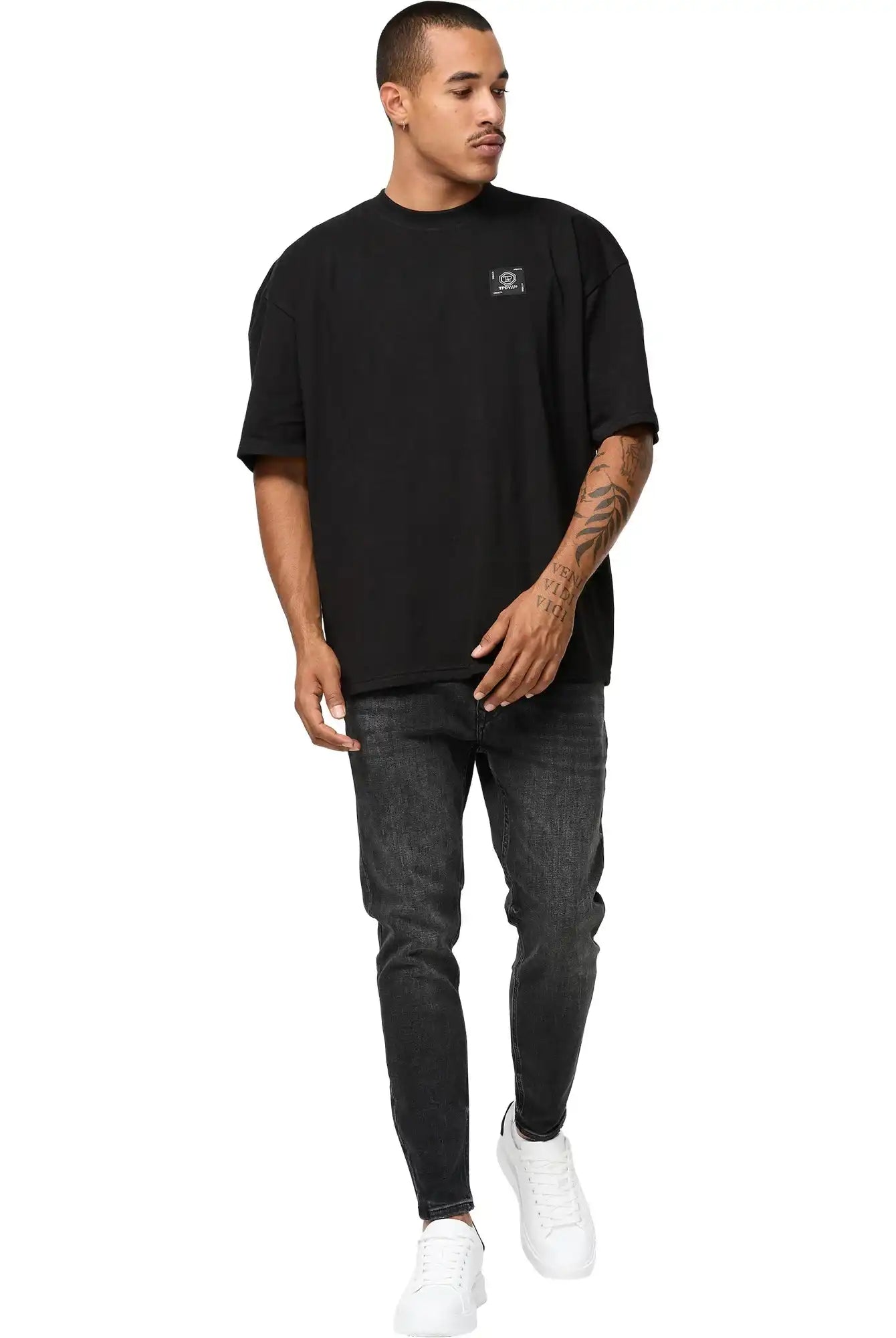 Ein Mann mit kurzen Haaren und einem Tattoo auf dem linken Unterarm trägt das trueprodigy Maxime Oversized T-Shirt mit Druck, gestylt als Unisex-T-Shirt, gepaart mit dunklen Jeans und weißen Turnschuhen. Er blickt zur Seite, während er vor einem weißen Hintergrund vorwärts geht.