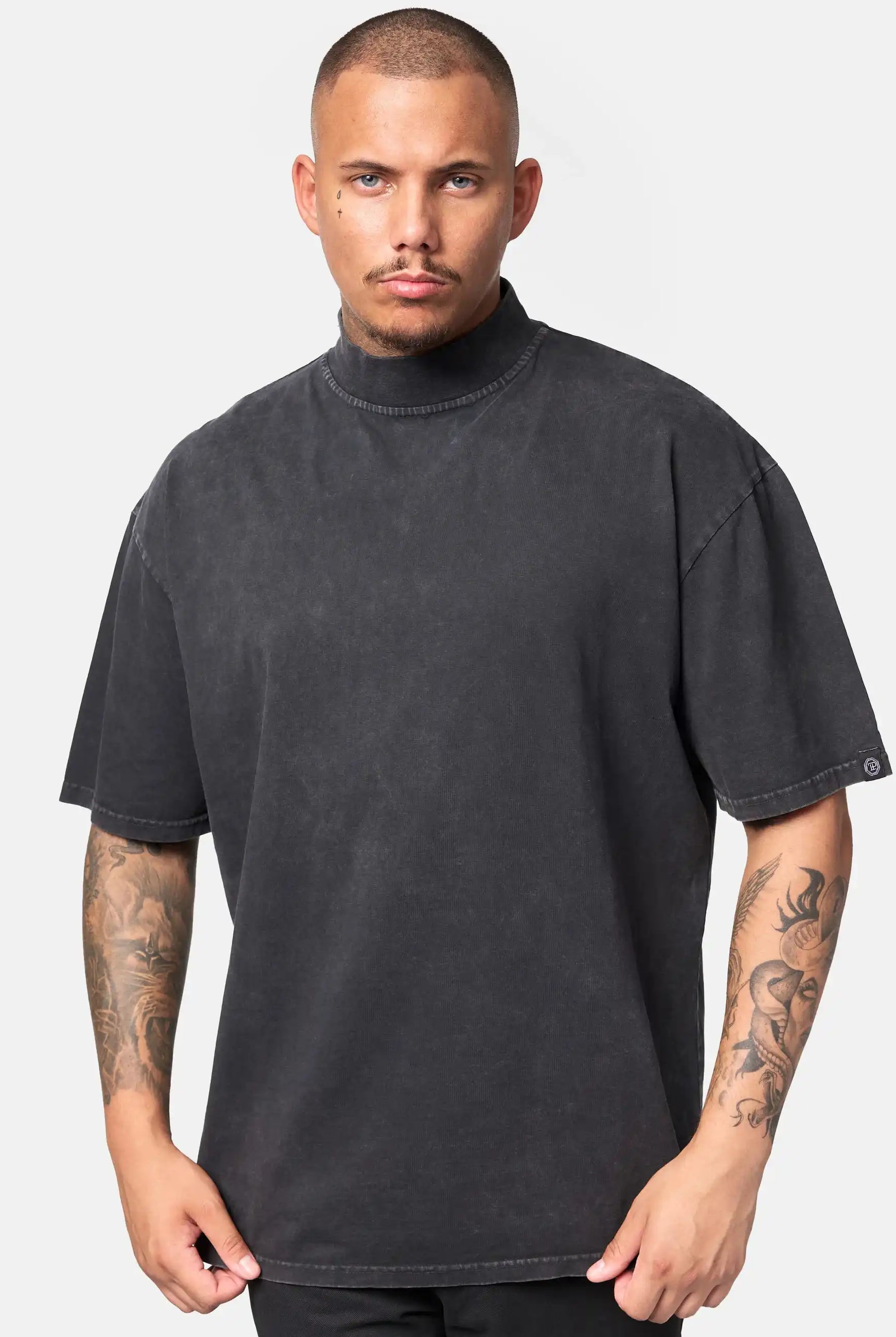 Ein Mann mit kurzen Haaren und sichtbaren Tattoos trägt das trueprodigy Phoenix Oversized T-Shirt mit Stehkragen in verblasstem Schwarz, gepaart mit einer schwarzen Hose, vor einem schlichten hellgrauen Hintergrund.