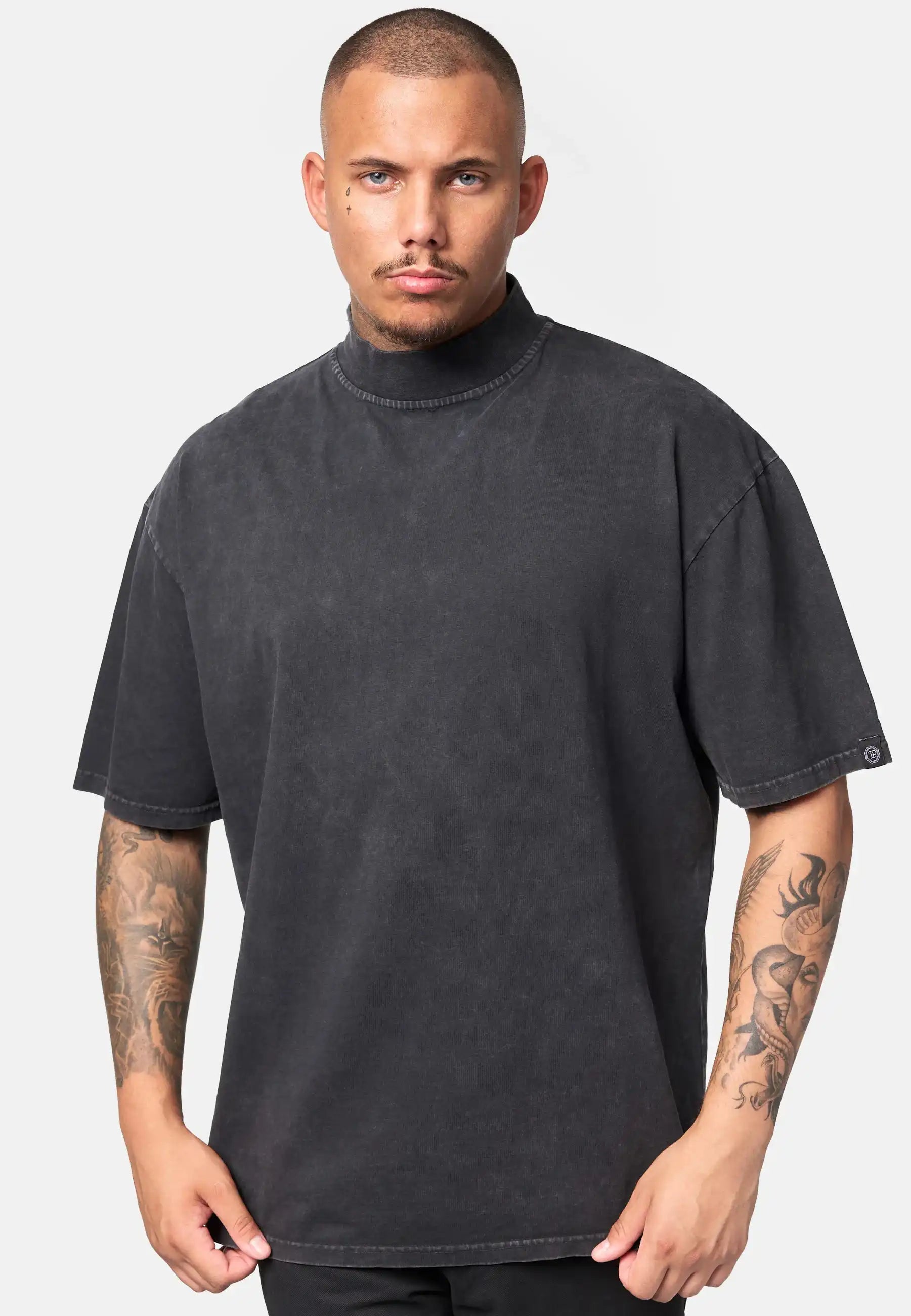Ein Mann mit kurzen Haaren und sichtbaren Tattoos trägt das trueprodigy Phoenix Oversized T-Shirt mit Stehkragen in verblasstem Schwarz, gepaart mit einer schwarzen Hose, vor einem schlichten hellgrauen Hintergrund.