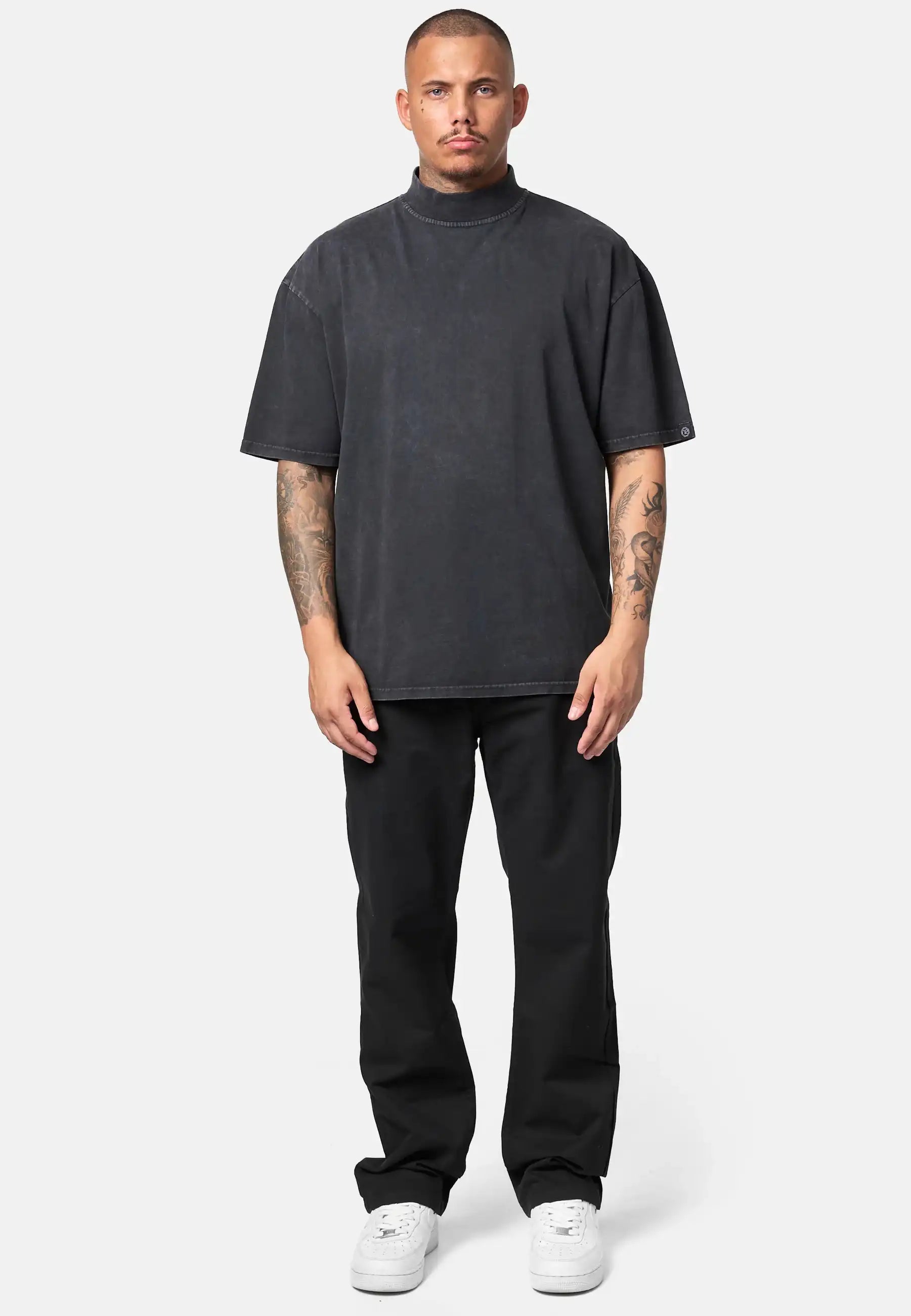 Ein Mann mit kurzen Haaren und Armtattoos steht mit dem Gesicht nach vorne und trägt das trueprodigy Phoenix Oversized T-Shirt mit Stehkragen, schwarze Hosen und weiße Turnschuhe vor einem schlichten hellgrauen Hintergrund.
