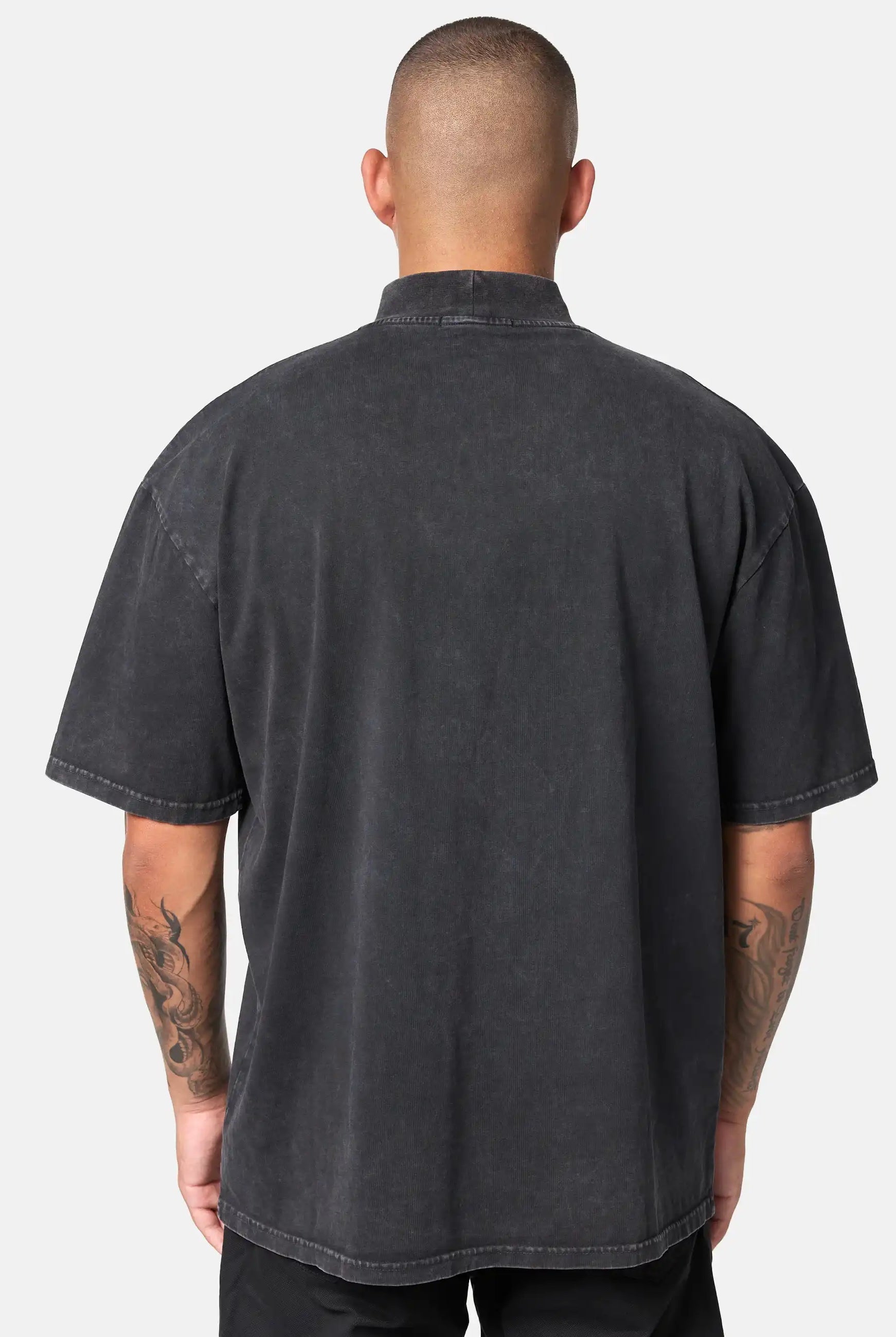 Man sieht eine Person mit kurzen Haaren von hinten, die das trueprodigy Phoenix Oversized T-Shirt mit Stehkragen in schwarz und schwarzer Hose trägt. Beide tätowierten Arme sind vor einem schlichten, hellen Hintergrund zu sehen.