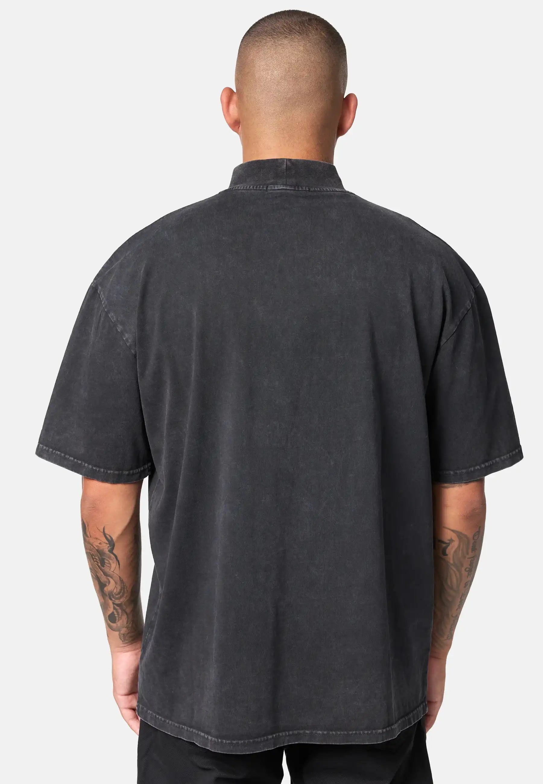 Man sieht eine Person mit kurzen Haaren von hinten, die das trueprodigy Phoenix Oversized T-Shirt mit Stehkragen in schwarz und schwarzer Hose trägt. Beide tätowierten Arme sind vor einem schlichten, hellen Hintergrund zu sehen.