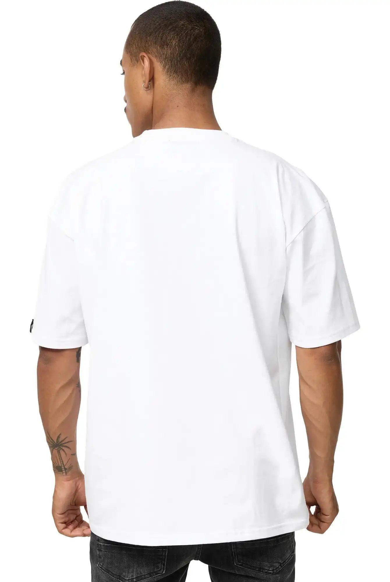 Eine Person mit kurzen Haaren und einer Tätowierung am linken Unterarm blickt von der Kamera weg und trägt ein schlichtes weißes trueprodigy Phoenix Oversized T-Shirt mit Stehkragen und dunkle Jeans.