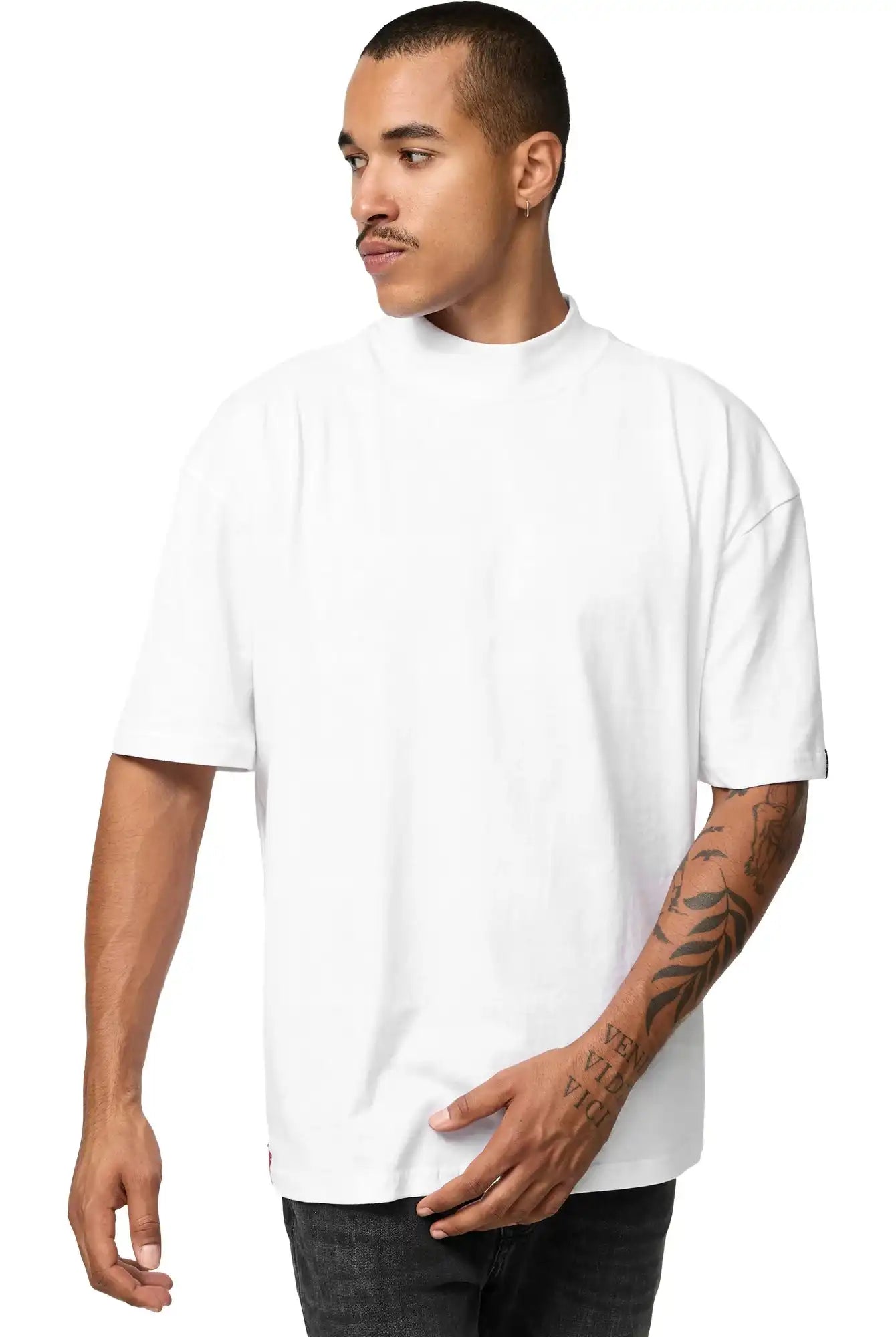 Ein Mann mit kurzen Haaren und Armtattoos trägt das trueprodigy Phoenix Oversized T-Shirt mit Stehkragen und schwarze Jeans. Er steht vor einem schlichten weißen Hintergrund und blickt mit neutraler Miene nach rechts.