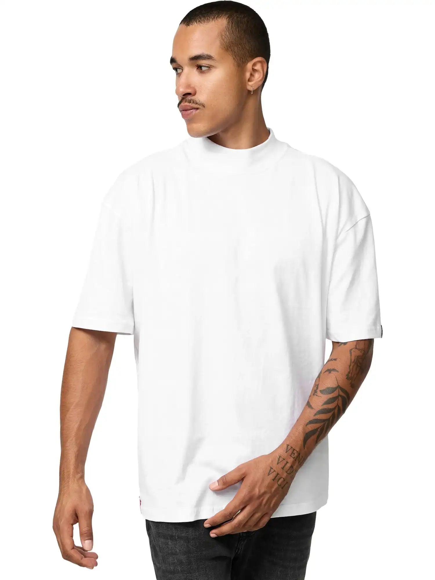 Ein Mann mit kurzen Haaren und Armtattoos trägt das trueprodigy Phoenix Oversized T-Shirt mit Stehkragen und schwarze Jeans. Er steht vor einem schlichten weißen Hintergrund und blickt mit neutraler Miene nach rechts.