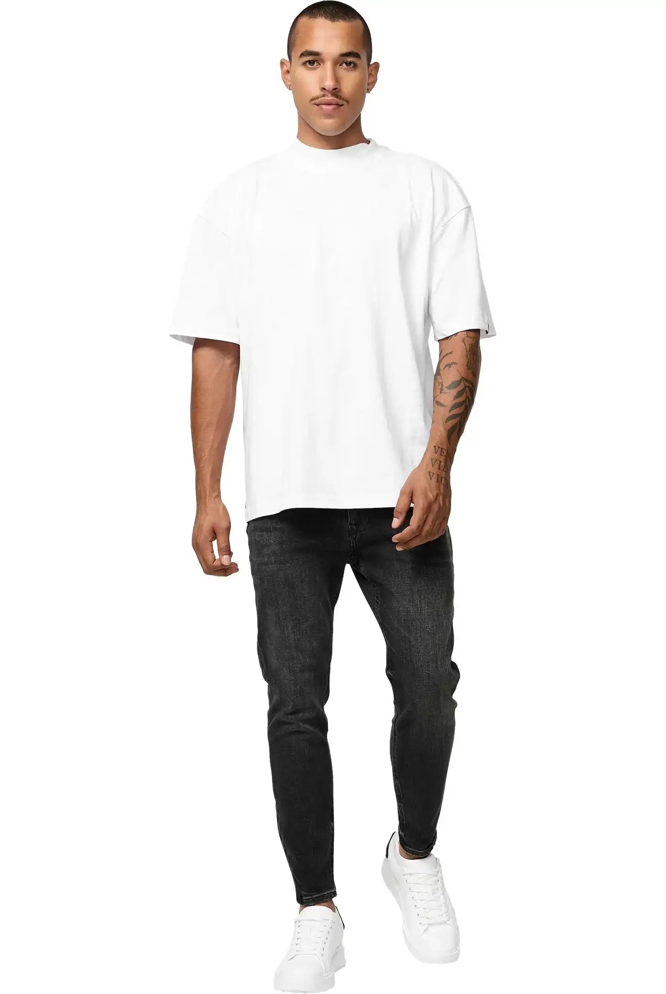 Ein Mann steht vor einem weißen Hintergrund und trägt das trueprodigy Phoenix Oversized T-Shirt mit Stehkragen in weiß, schwarze Slim-Fit Jeans und weiße Turnschuhe. Seine rechte Hand liegt an seiner Seite und sein linker Arm ist tätowiert.