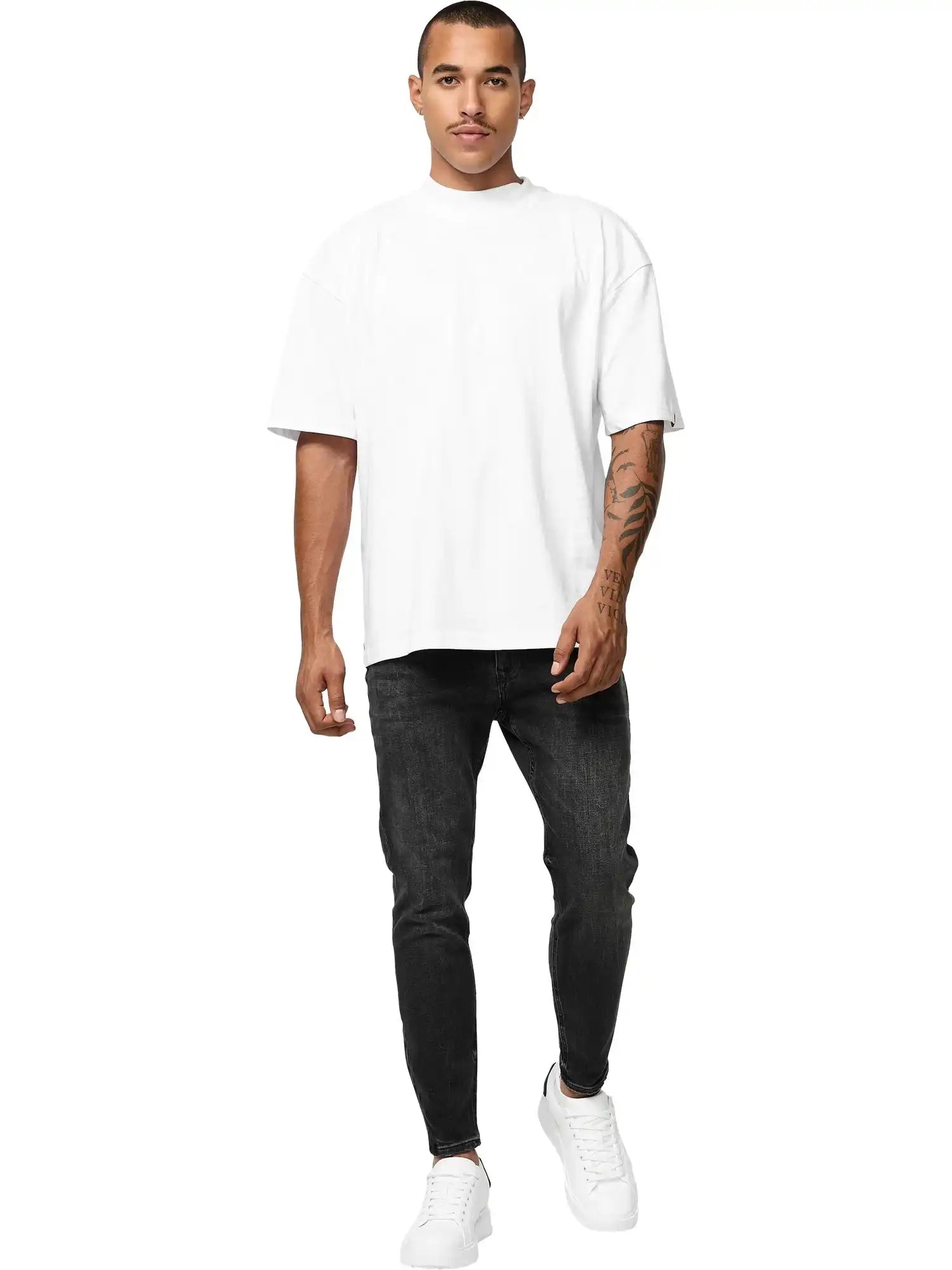 Ein Mann steht vor einem weißen Hintergrund und trägt das trueprodigy Phoenix Oversized T-Shirt mit Stehkragen in weiß, schwarze Slim-Fit Jeans und weiße Turnschuhe. Seine rechte Hand liegt an seiner Seite und sein linker Arm ist tätowiert.