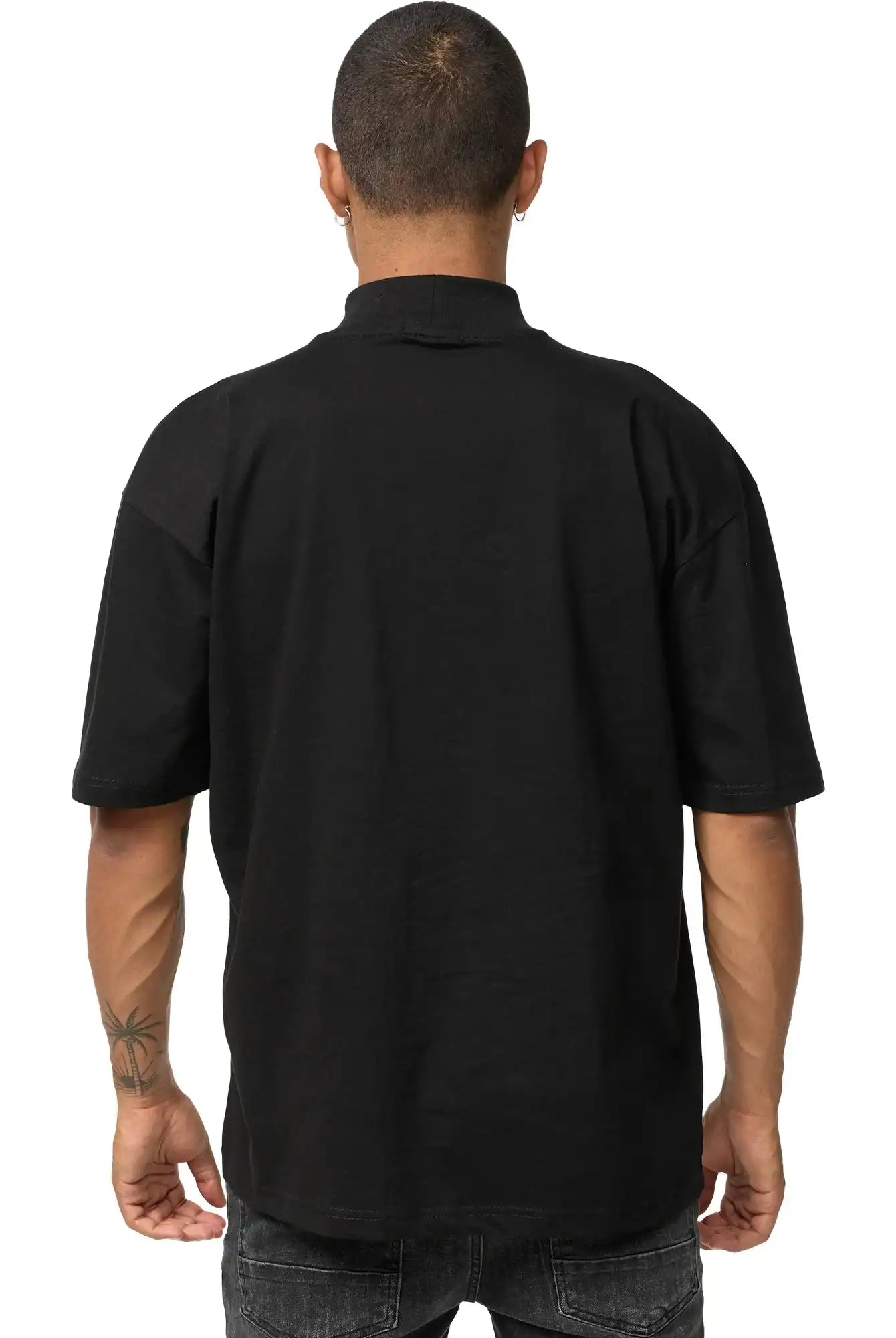 Eine Person mit kurz geschnittenem Haar blickt von der Kamera weg und trägt das schwarze Phoenix Oversized T-Shirt mit Stehkragen von trueprodigy und dunkle Jeans. Ihre Arme sind sichtbar und auf ihrem linken Unterarm ist ein Tattoo zu sehen.