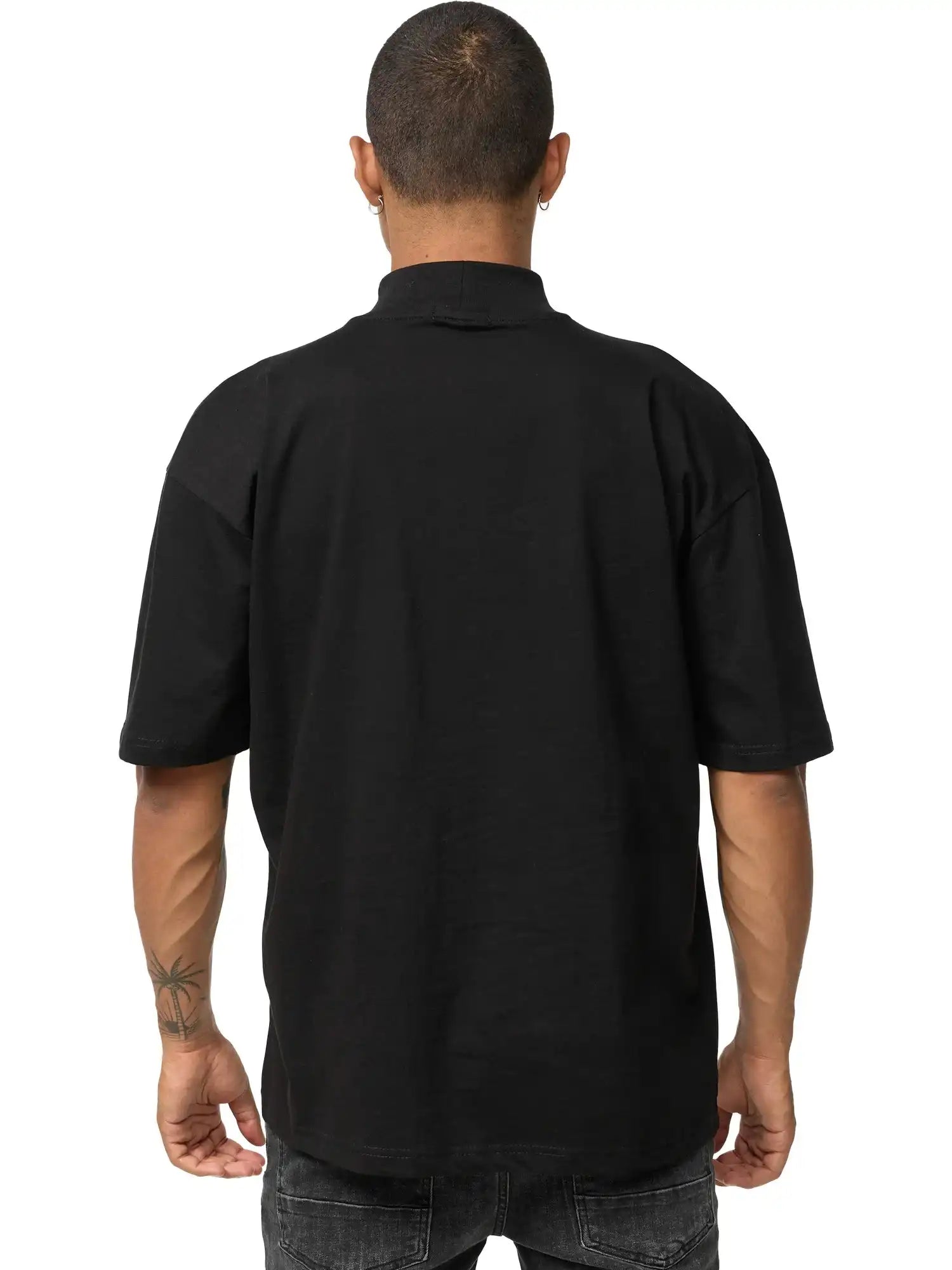 Eine Person mit kurz geschnittenem Haar blickt von der Kamera weg und trägt das schwarze Phoenix Oversized T-Shirt mit Stehkragen von trueprodigy und dunkle Jeans. Ihre Arme sind sichtbar und auf ihrem linken Unterarm ist ein Tattoo zu sehen.