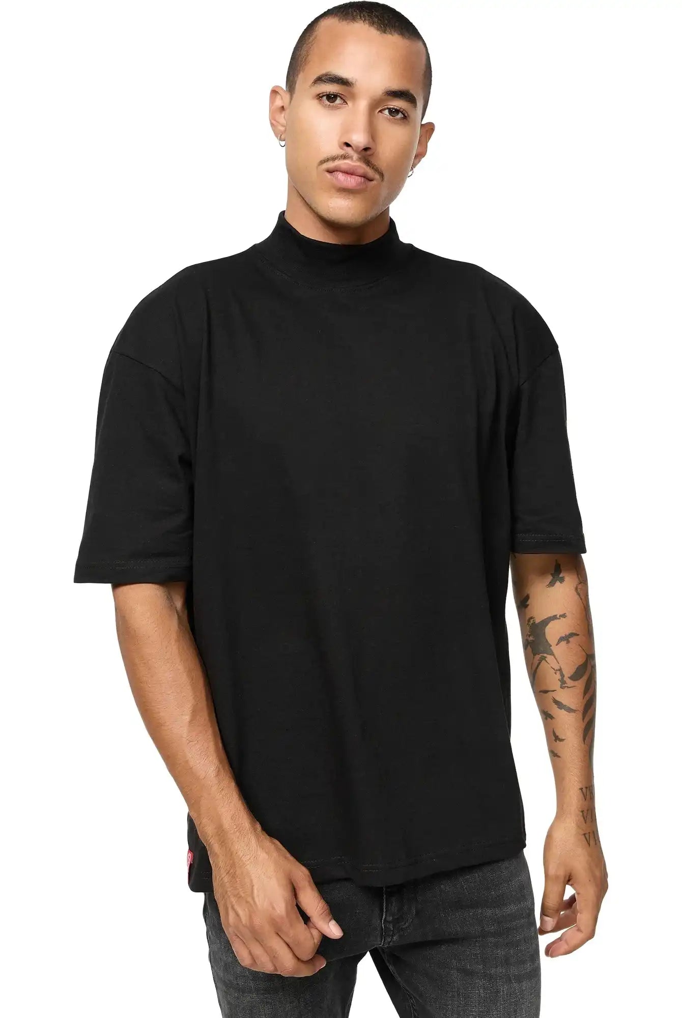 Eine Person mit kurzen Haaren und einem Schnurrbart trägt das trueprodigy Phoenix Oversized T-Shirt mit Stehkragen in schwarz und dunkler Jeans, steht mit angewinkeltem Arm vor einem schlichten weißen Hintergrund. Auf ihrem linken Unterarm sind Tätowierungen zu sehen.