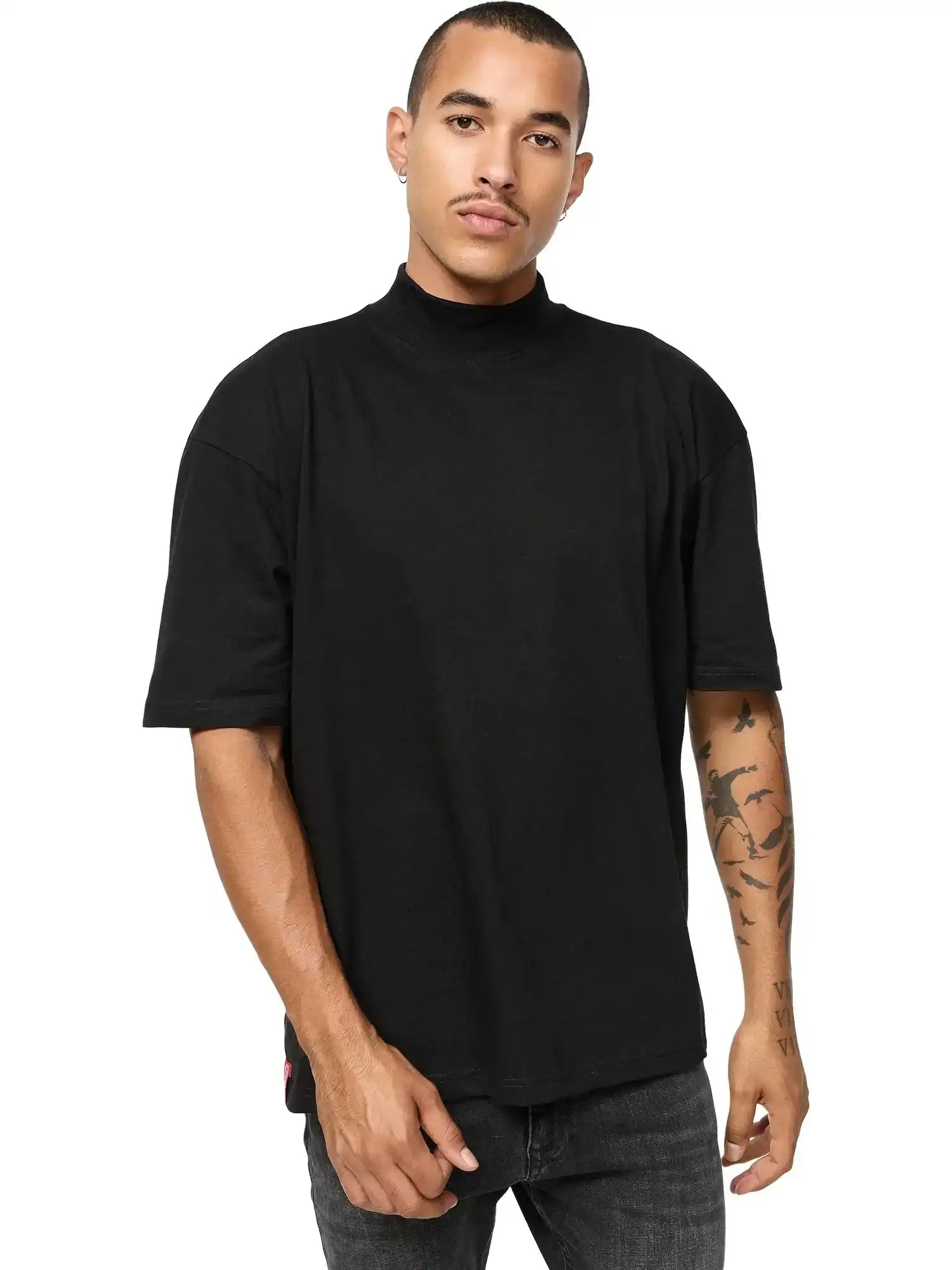 Eine Person mit kurzen Haaren und einem Schnurrbart trägt das trueprodigy Phoenix Oversized T-Shirt mit Stehkragen in schwarz und dunkler Jeans, steht mit angewinkeltem Arm vor einem schlichten weißen Hintergrund. Auf ihrem linken Unterarm sind Tätowierungen zu sehen.