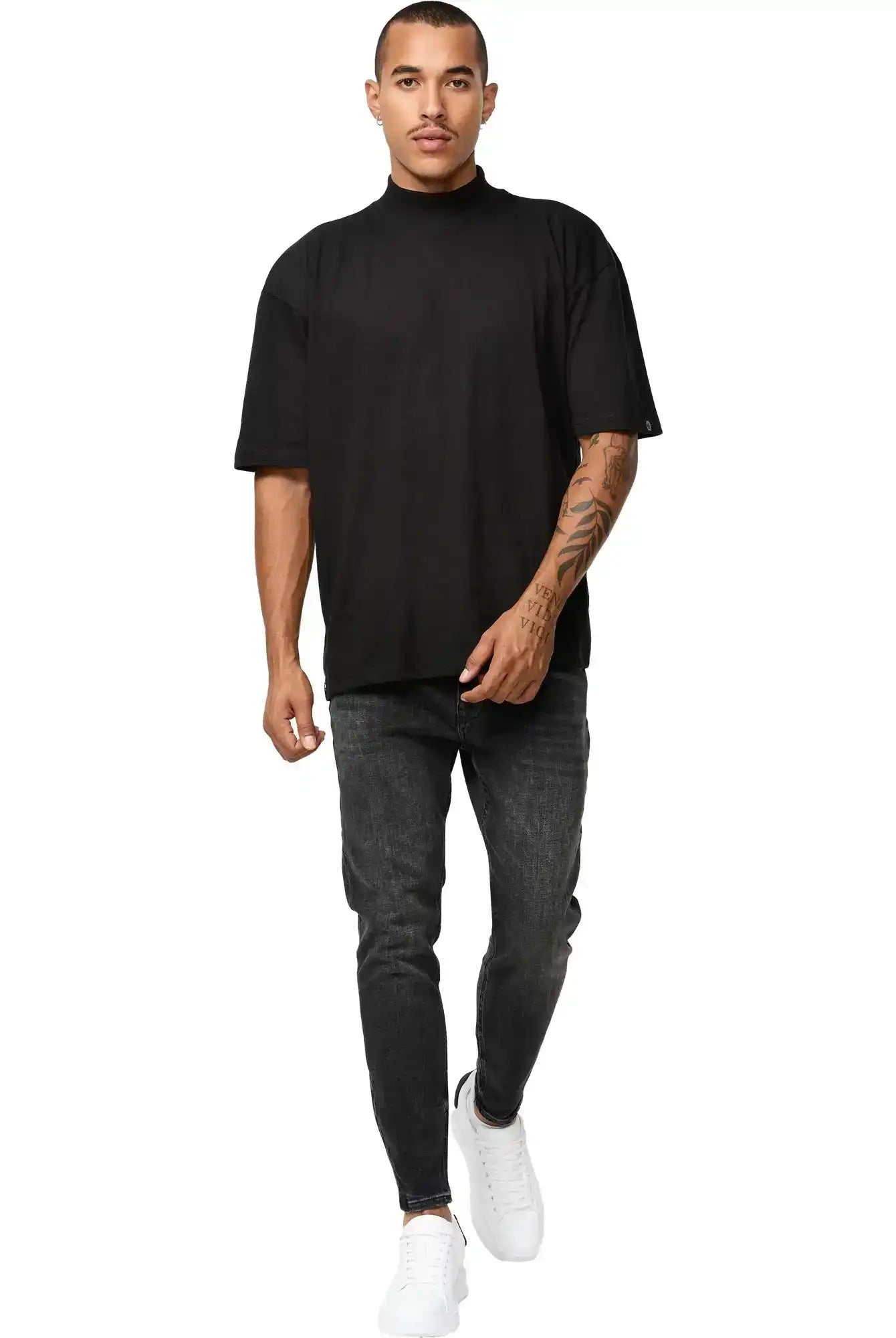 Ein Mann mit kurzen Haaren und einem sichtbaren Tattoo auf dem rechten Arm geht mit neutraler Miene nach vorne. Er trägt das trueprodigy Phoenix Oversized T-Shirt mit Stehkragen, dunkelgraue Jeans und weiße Turnschuhe vor einem schlichten weißen Hintergrund.