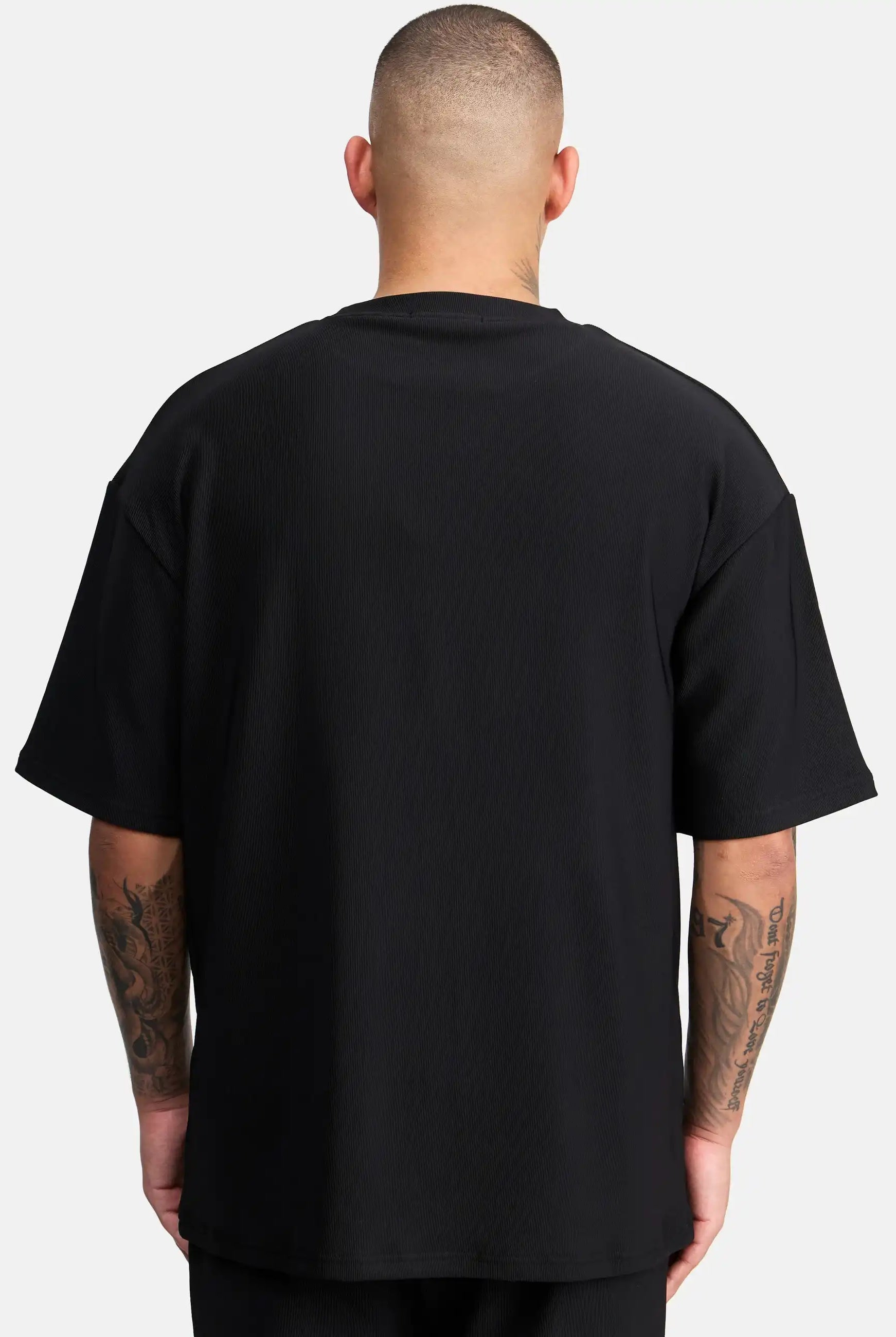 Eine tätowierte Person mit rasiertem Kopf ist von hinten zu sehen, die das trueprodigy Mateo Oversized T-Shirt mit Stickerei in schwarz und kurzen Ärmeln trägt und vor einem schlichten weißen Hintergrund steht.