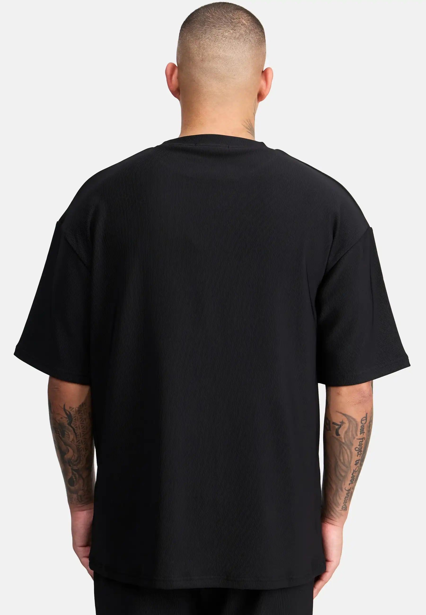 Eine tätowierte Person mit rasiertem Kopf ist von hinten zu sehen, die das trueprodigy Mateo Oversized T-Shirt mit Stickerei in schwarz und kurzen Ärmeln trägt und vor einem schlichten weißen Hintergrund steht.