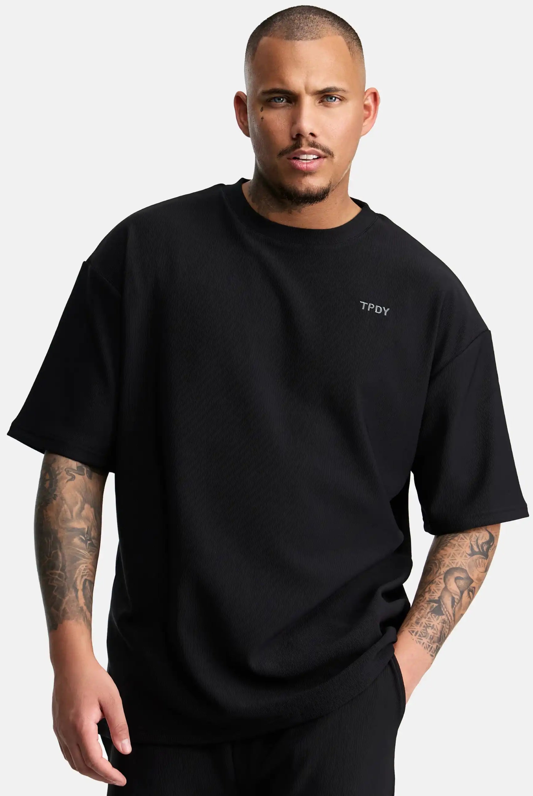 Ein Mann mit kurzen Haaren und Tattoos auf beiden Armen trägt das trueprodigy Mateo Oversized T-Shirt mit Stickerei. Er steht vor einem schlichten weißen Hintergrund und schaut in die Kamera.