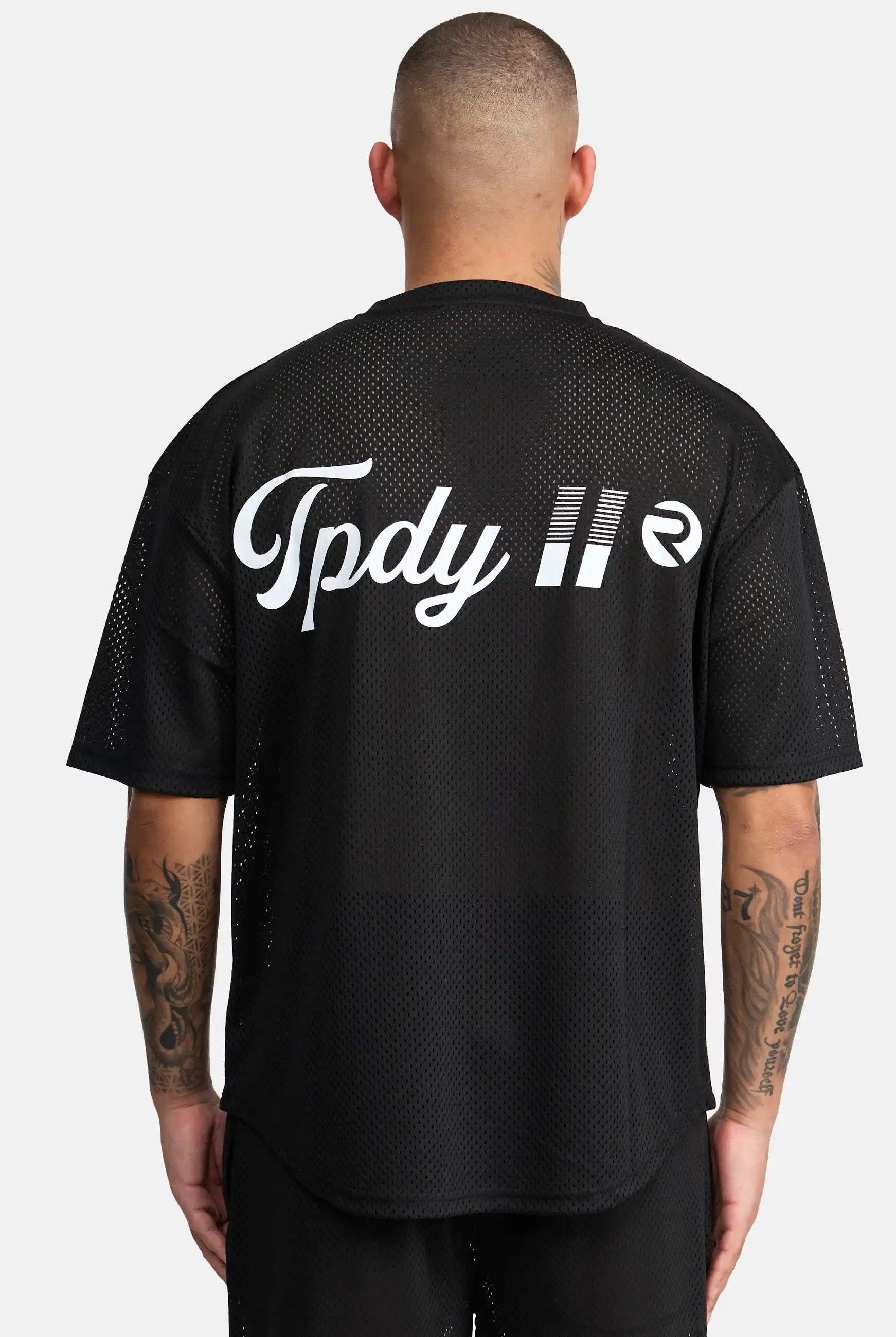 Eine Person mit kurzen Haaren und tätowierten Armen blickt in die Ferne. Sie trägt das trueprodigy Daniel Oversized T-Shirt mit Druck in schwarzem Mesh mit dem Schriftzug "Tpdy II" und einem kreisförmigen weißen Logo auf dem Rücken vor einem schlichten weißen Hintergrund.
