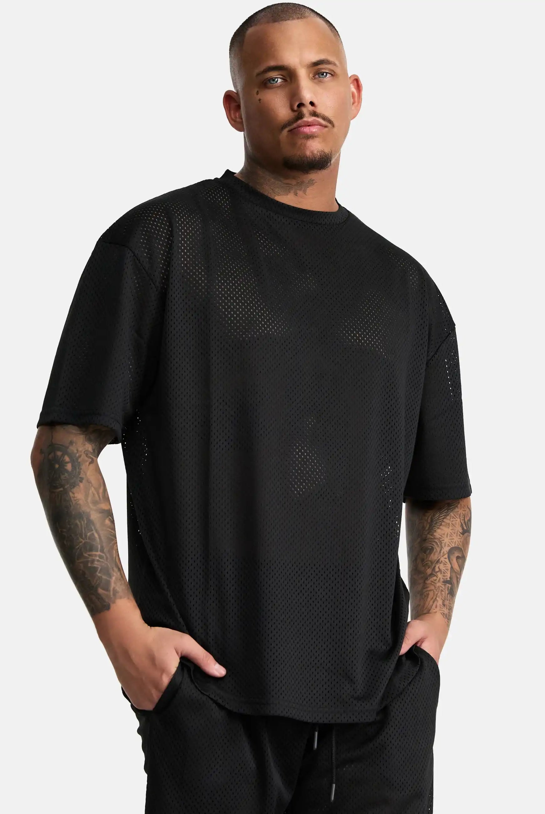Ein Mann mit kurzen Haaren und sichtbaren Arm-Tattoos trägt das Daniel Oversized T-Shirt mit Druck von trueprodigy in schwarz, gepaart mit einer schwarzen Hose, die Hände in den Taschen vor einem schlichten weißen Hintergrund für Vintage-Vibes.