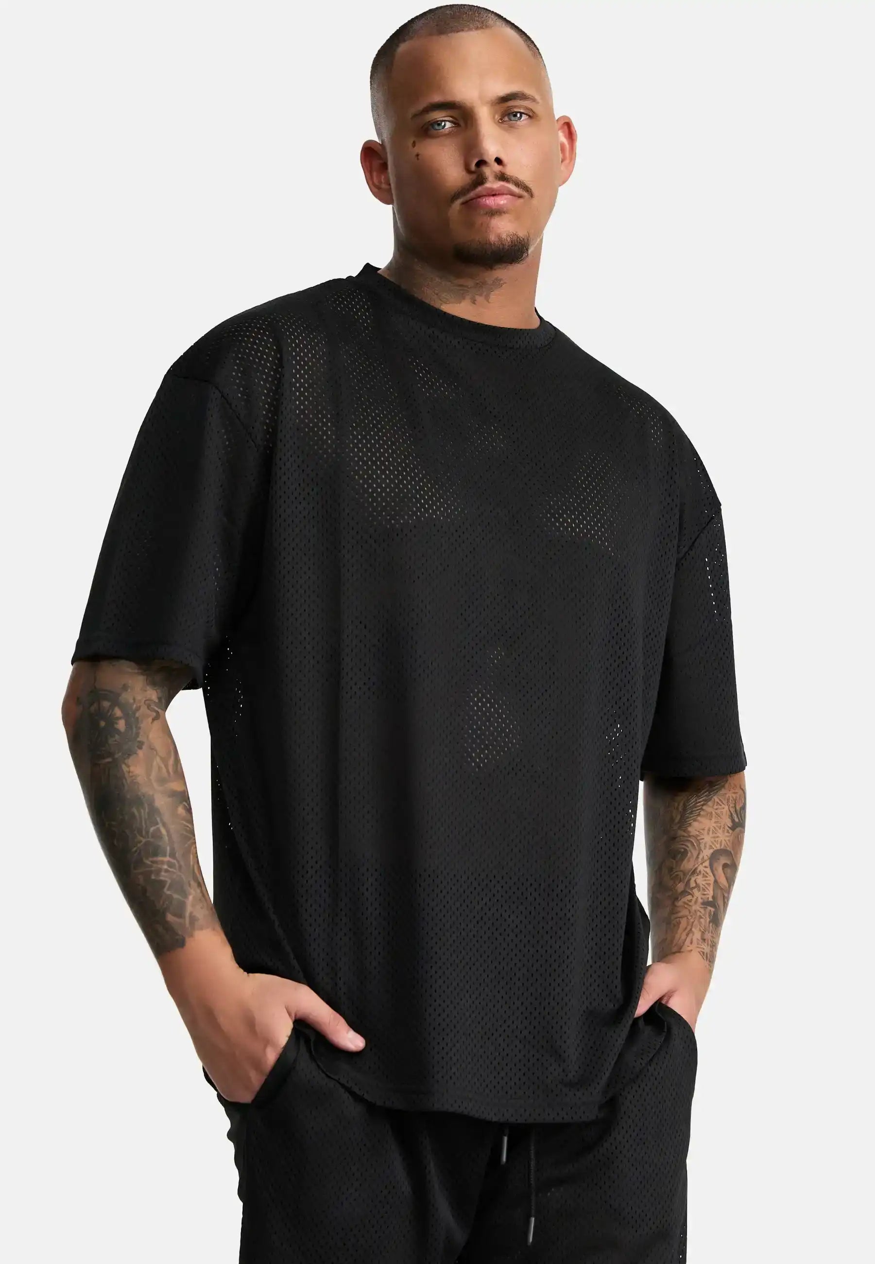 Ein Mann mit kurzen Haaren und sichtbaren Arm-Tattoos trägt das Daniel Oversized T-Shirt mit Druck von trueprodigy in schwarz, gepaart mit einer schwarzen Hose, die Hände in den Taschen vor einem schlichten weißen Hintergrund für Vintage-Vibes.