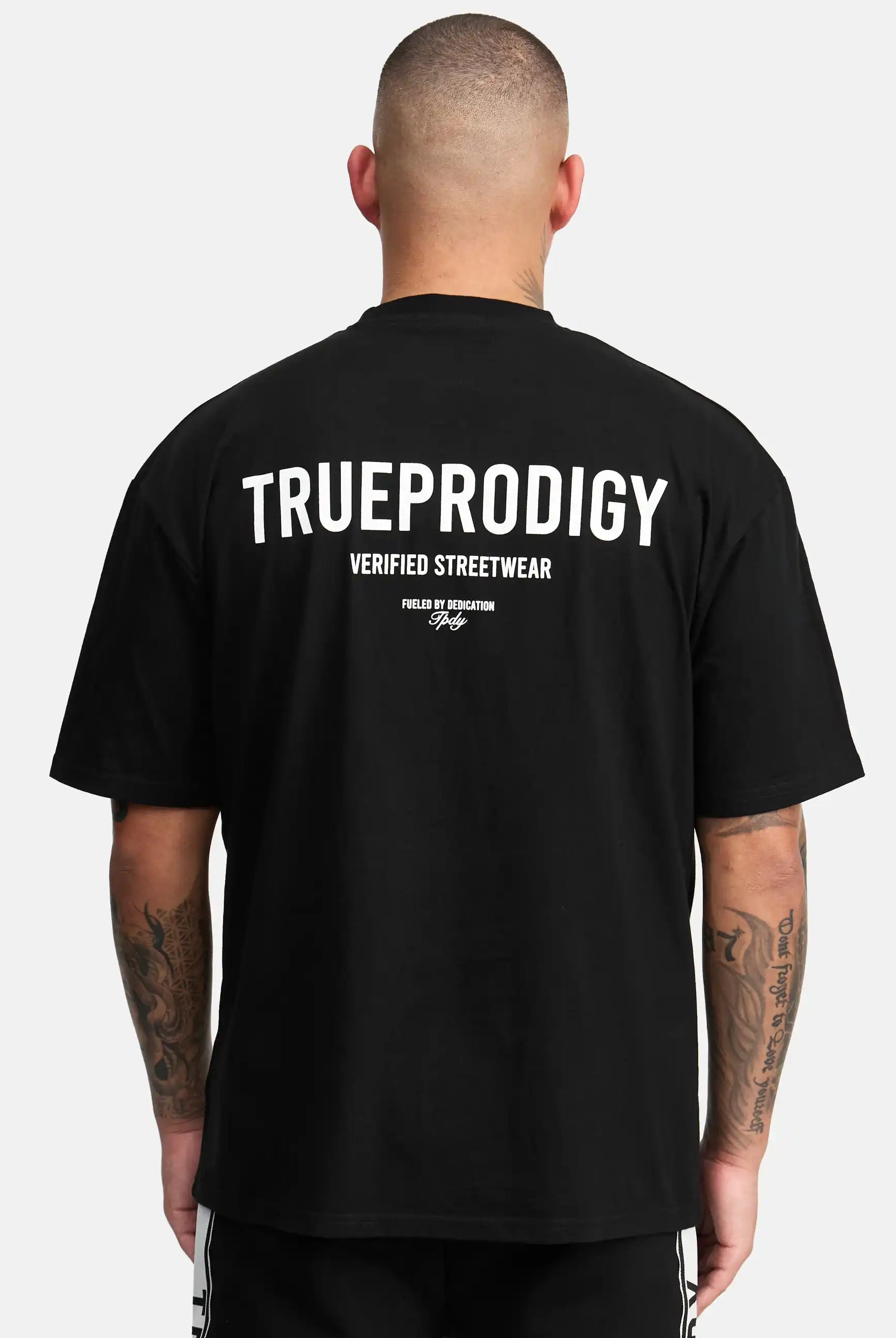 Eine Person mit kurzen Haaren und Tattoos auf beiden Unterarmen wird von hinten gezeigt. Sie trägt das trueprodigy Oliver Oversized T-Shirt mit Druck in schwarz, mit weißem "TRUEPRODIGY VERIFIED STREETWEAR" Aufdruck.