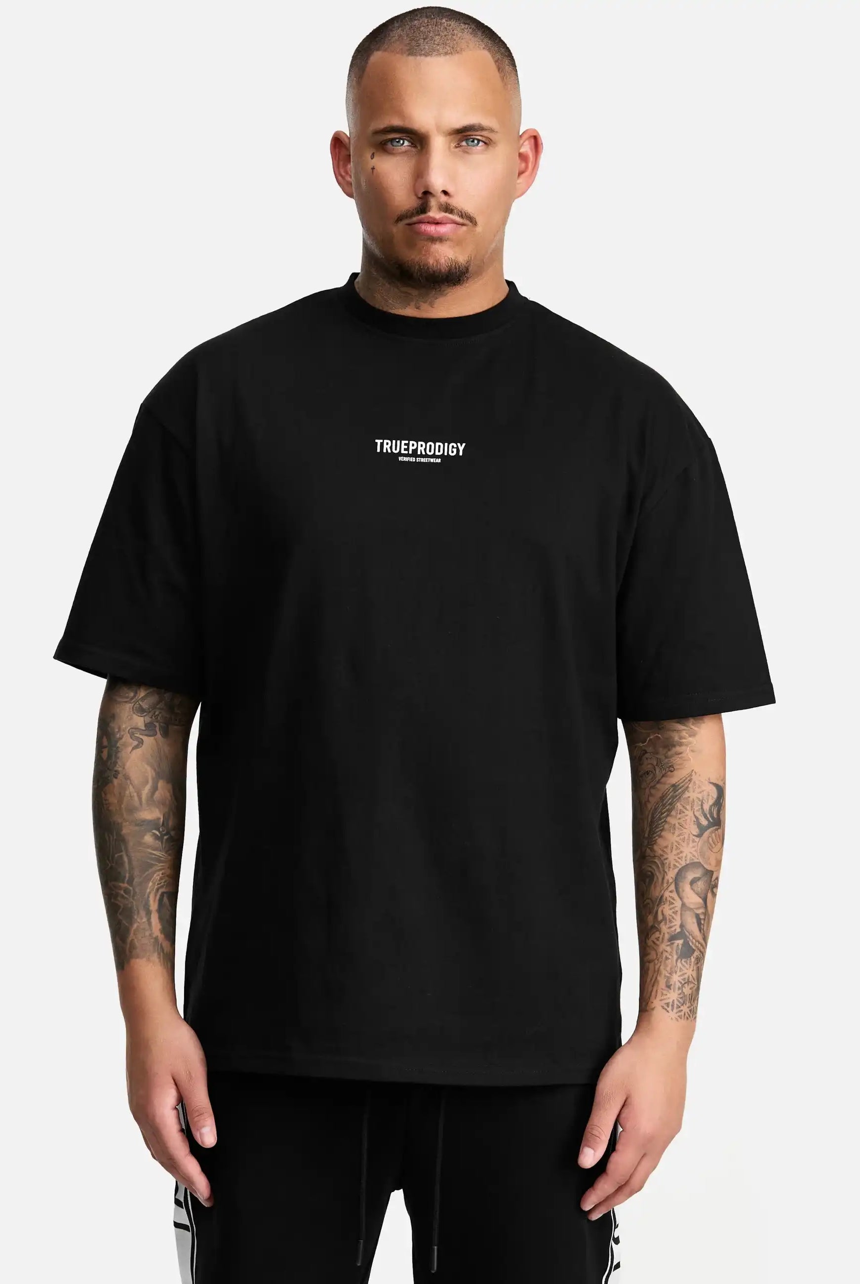 Ein tätowierter Mann blickt mit neutralem Blick nach vorne und trägt das trueprodigy Oliver Oversized T-Shirt mit Druck aus schwerem Jersey. Das lockere Unisex-Shirt zeigt das weiße Logo der Marke auf der Brust vor einem schlichten weißen Hintergrund.