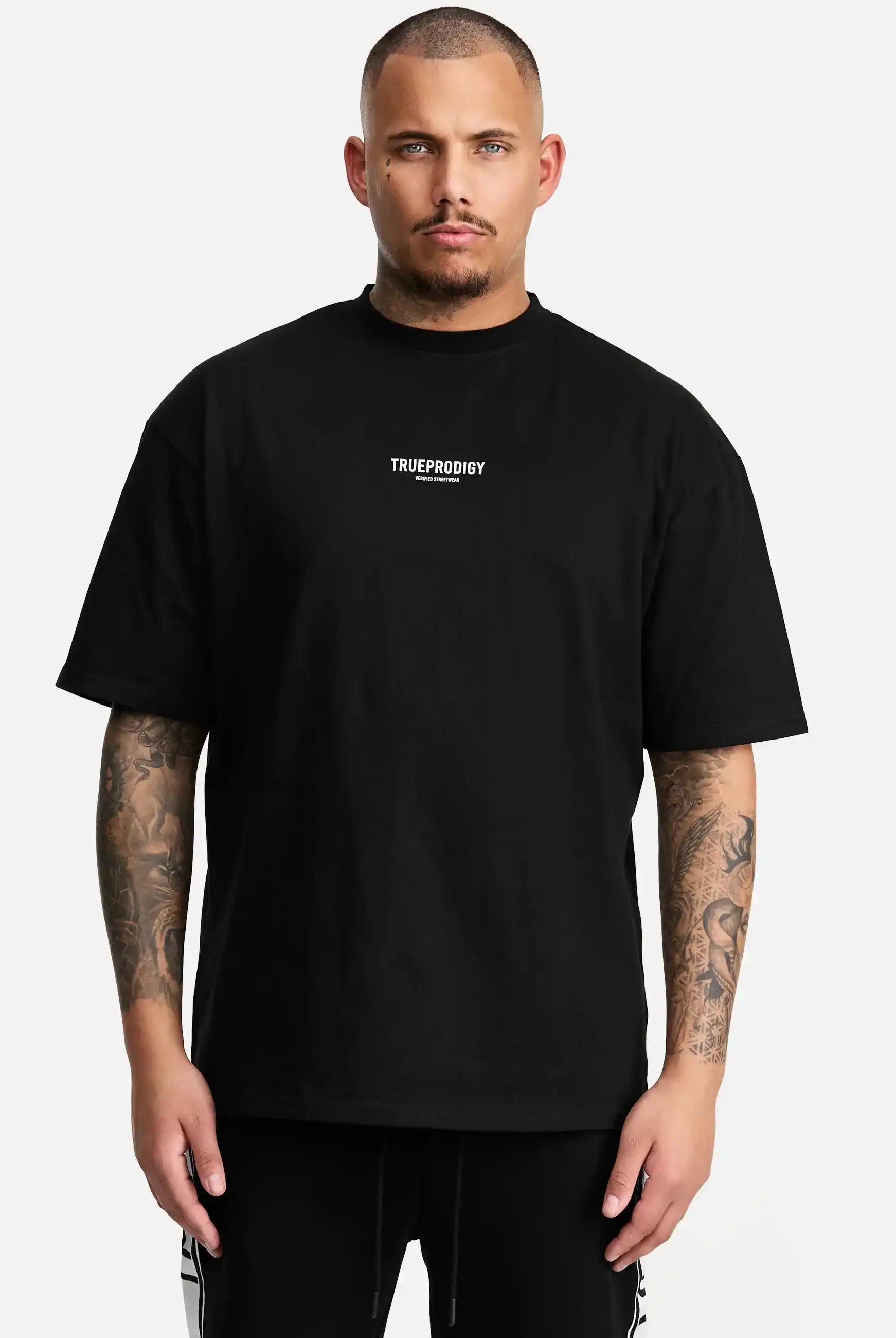 Ein tätowierter Mann blickt mit neutralem Blick nach vorne und trägt das trueprodigy Oliver Oversized T-Shirt mit Druck aus schwerem Jersey. Das lockere Unisex-Shirt zeigt das weiße Logo der Marke auf der Brust vor einem schlichten weißen Hintergrund.