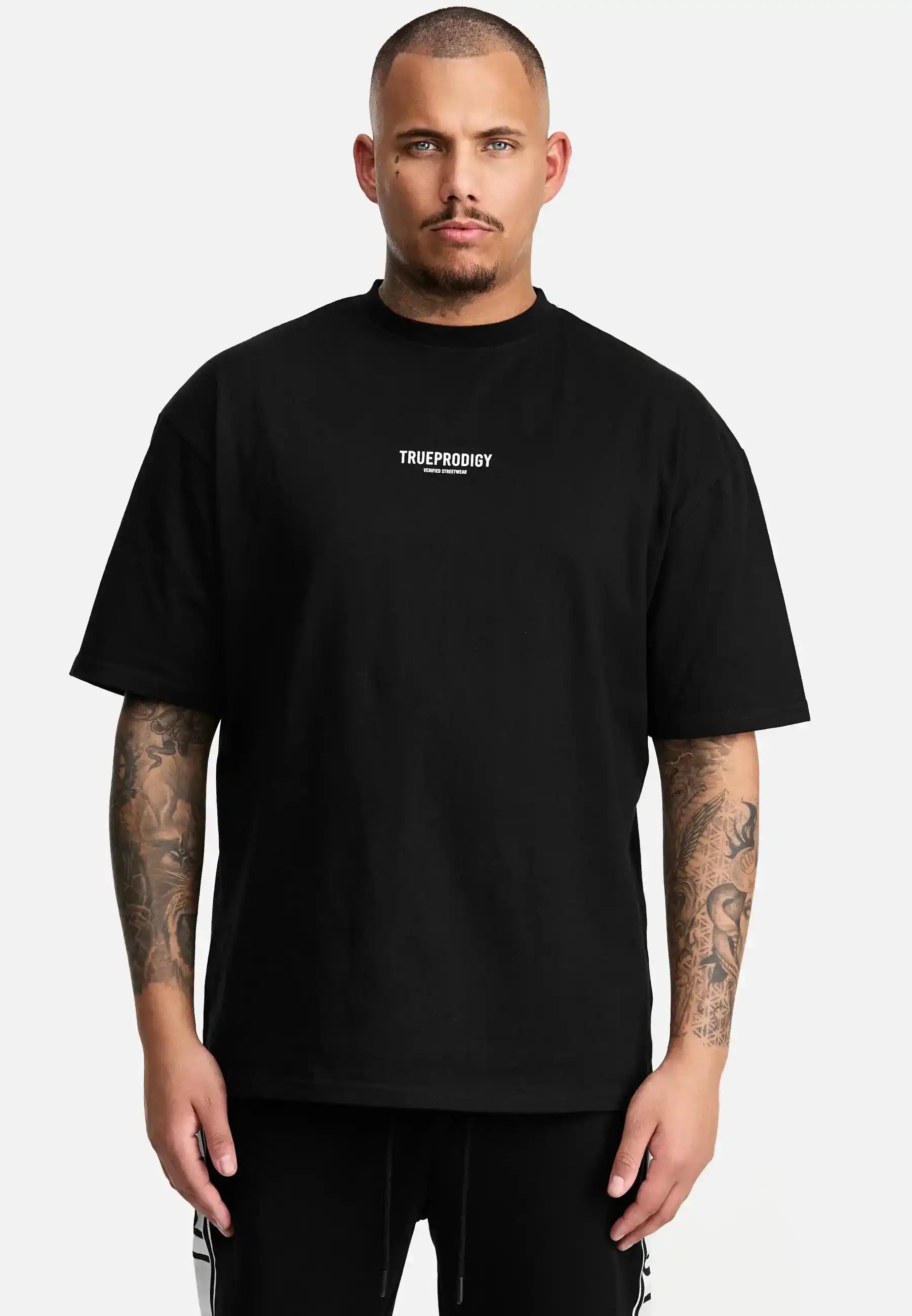 Ein tätowierter Mann blickt mit neutralem Blick nach vorne und trägt das trueprodigy Oliver Oversized T-Shirt mit Druck aus schwerem Jersey. Das lockere Unisex-Shirt zeigt das weiße Logo der Marke auf der Brust vor einem schlichten weißen Hintergrund.