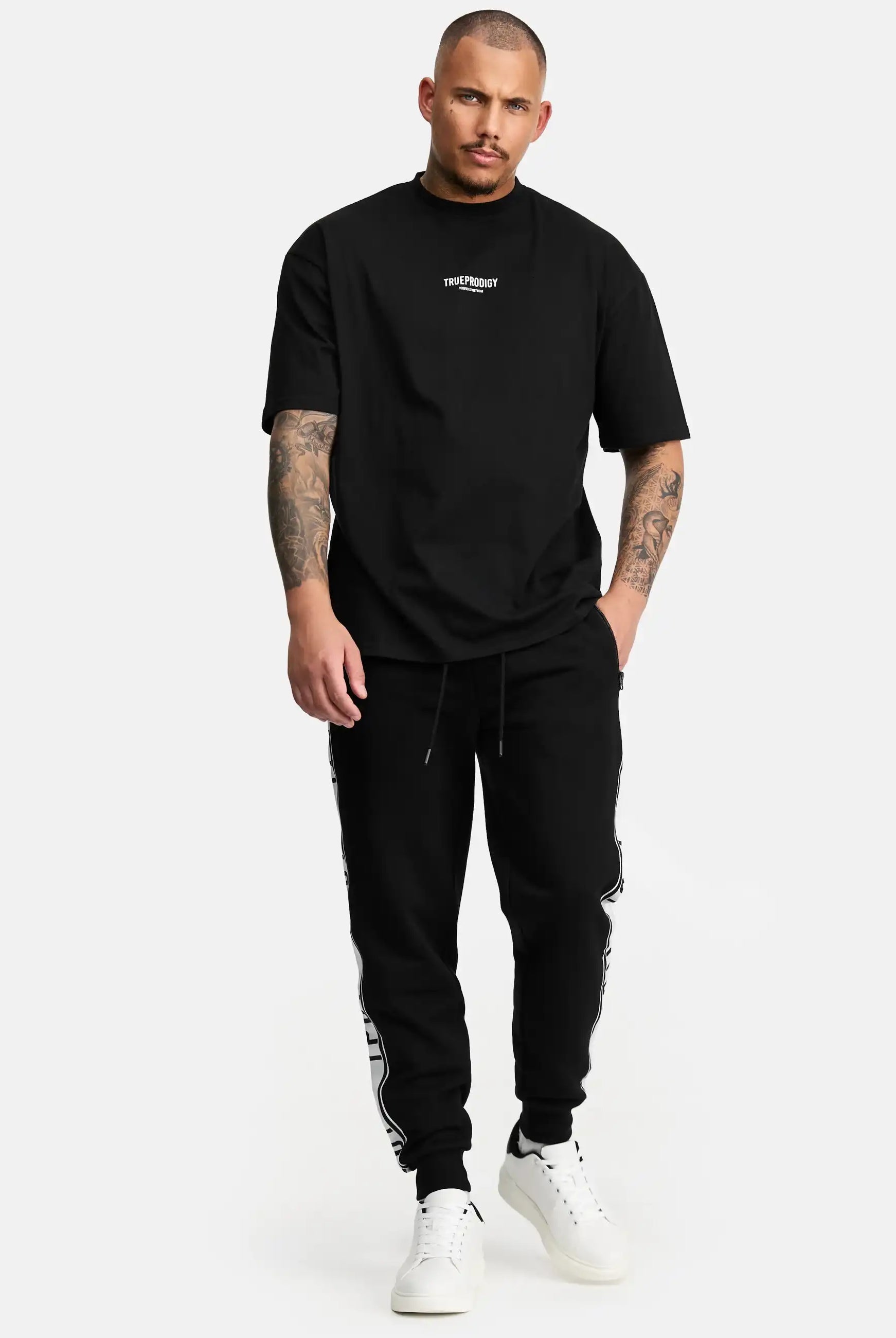 Ein Mann mit kurzen Haaren und Tattoos trägt das trueprodigy Oliver Oversized T-Shirt mit Druck, gepaart mit einer schwarzen Jogginghose mit weißen Seitendetails und weißen Turnschuhen, vor einem schlichten weißen Hintergrund.
