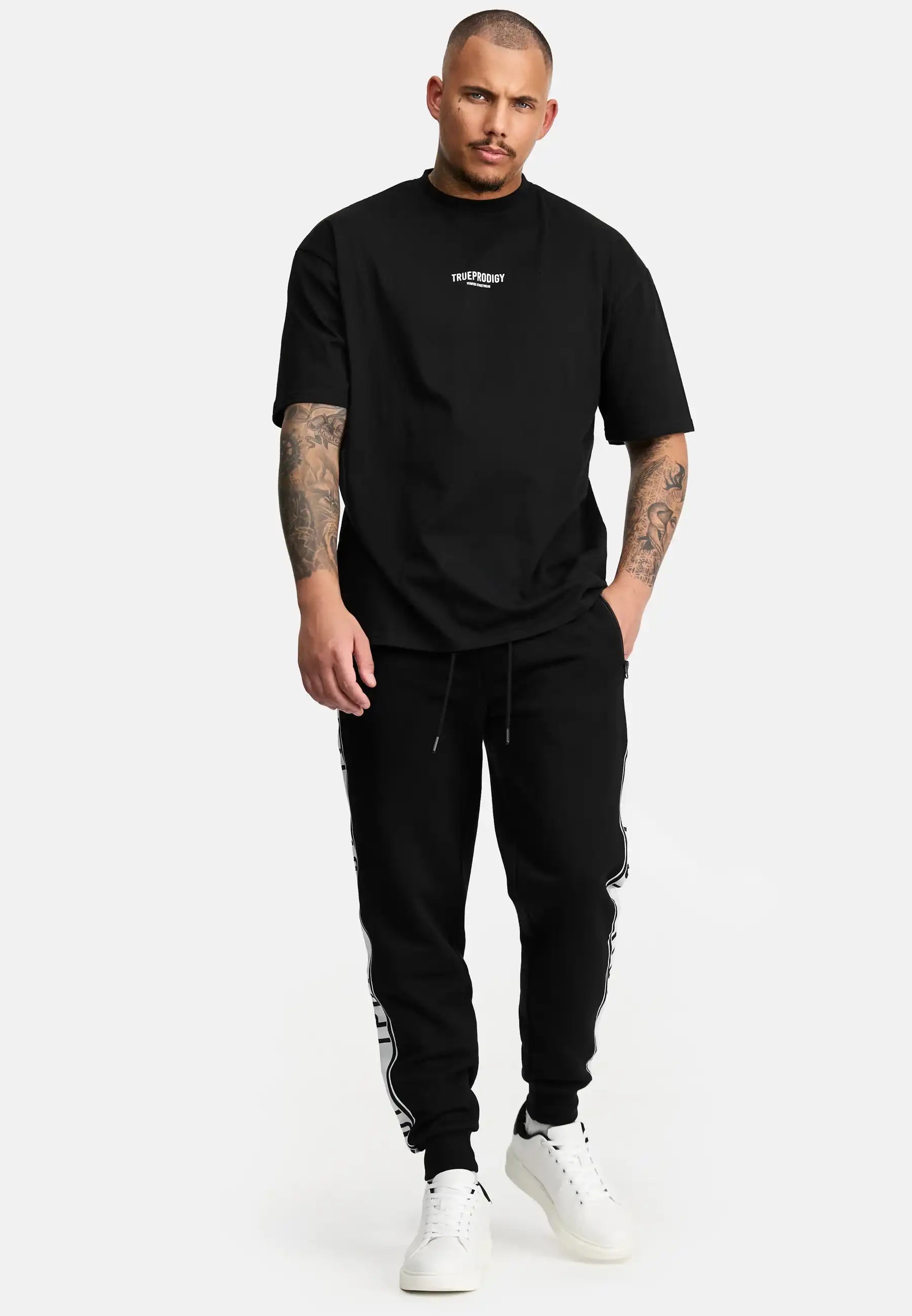 Ein Mann mit kurzen Haaren und Tattoos trägt das trueprodigy Oliver Oversized T-Shirt mit Druck, gepaart mit einer schwarzen Jogginghose mit weißen Seitendetails und weißen Turnschuhen, vor einem schlichten weißen Hintergrund.