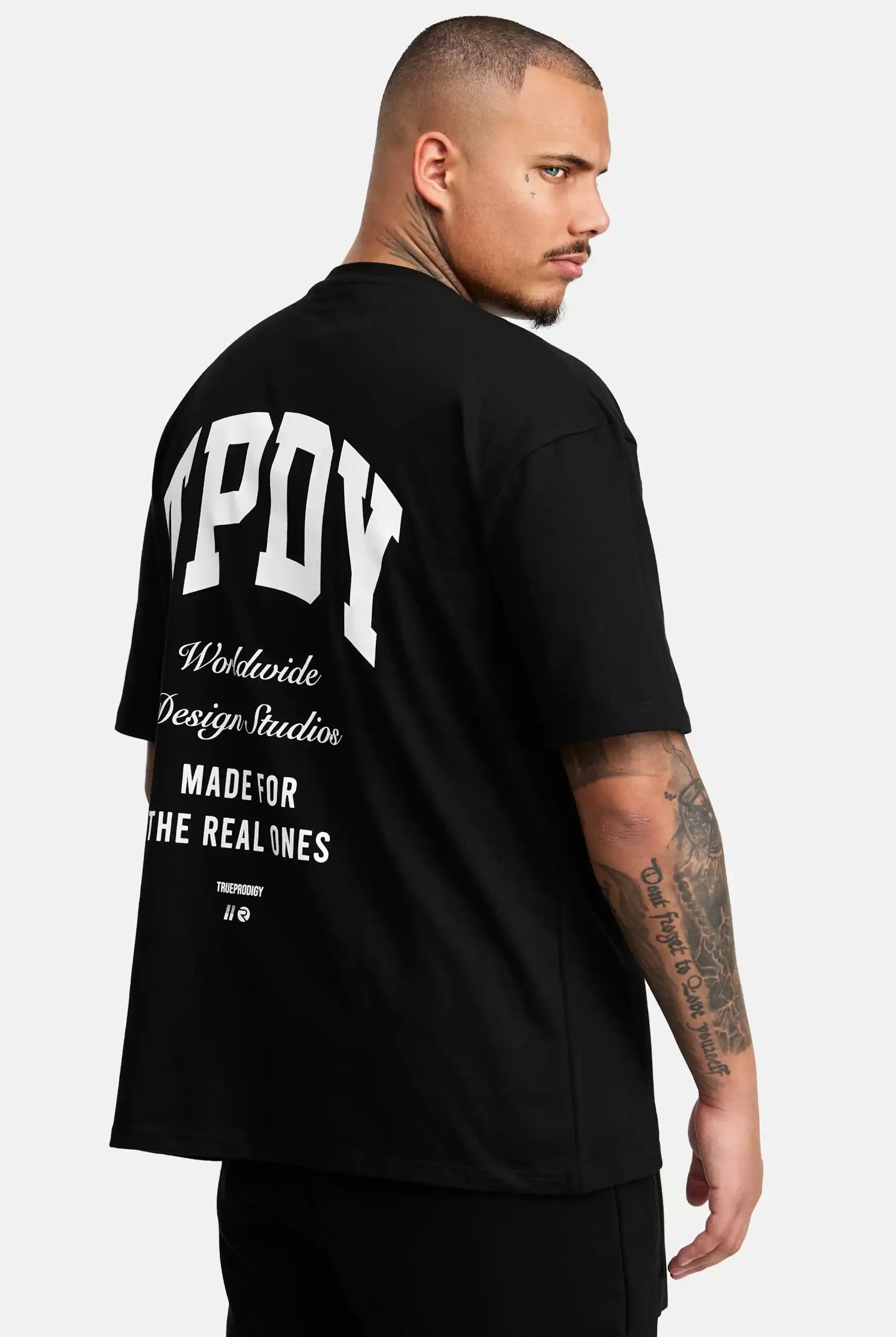 Ein Mann mit kurzen Haaren und Tattoos auf dem rechten Arm trägt das schwarze Elijah Oversized T-Shirt mit Druck von trueprodigy, mit weißem "MPOY Worldwide Design Studio Made for the Real Ones"-Text auf der Rückseite, und schaut über seine linke Schulter.